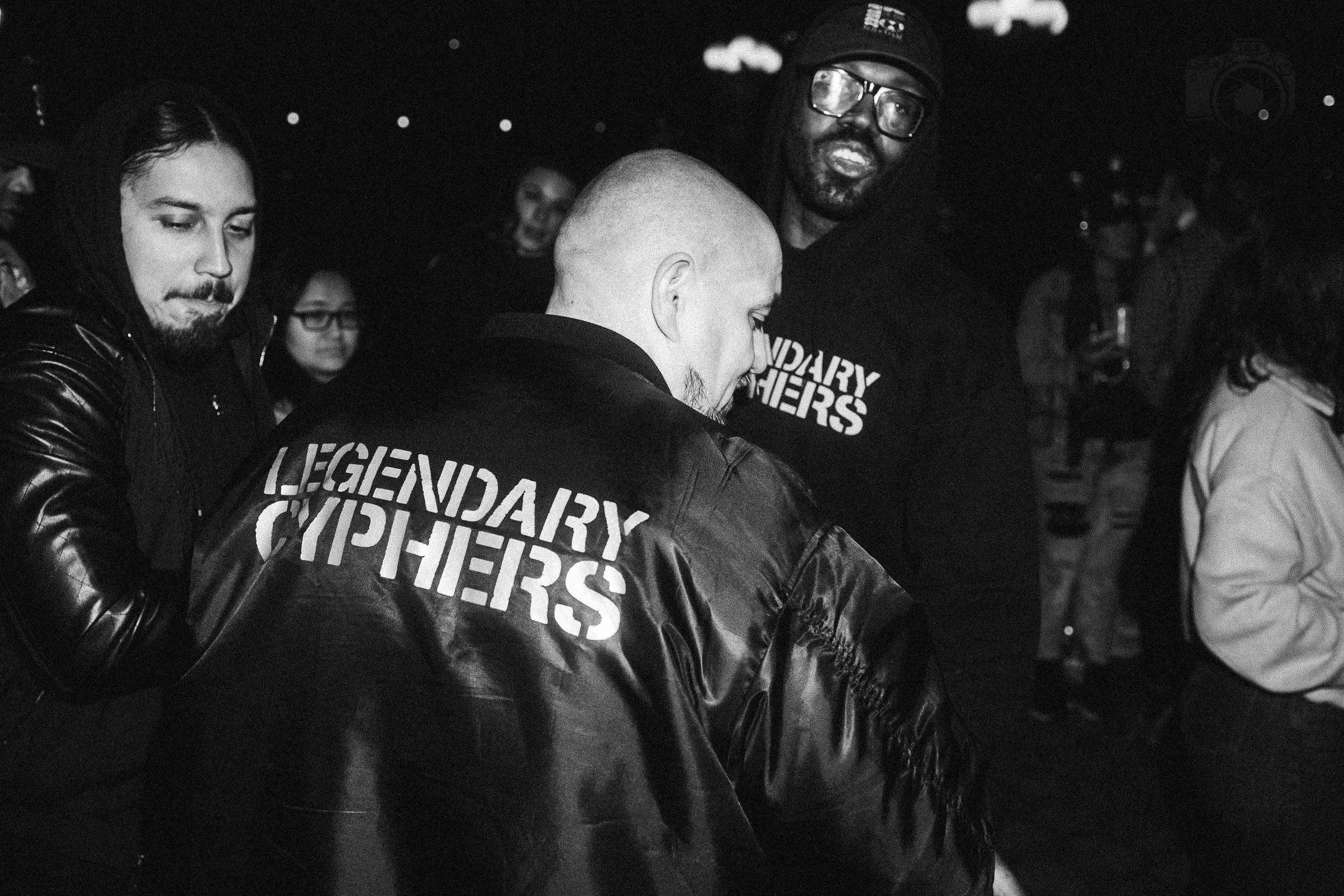 Group of people at a dark indoor event, some wearing jackets with 'LEGENDARY CYCLISTS' written on the back. One person has a shaved head and another is wearing glasses and a cap.