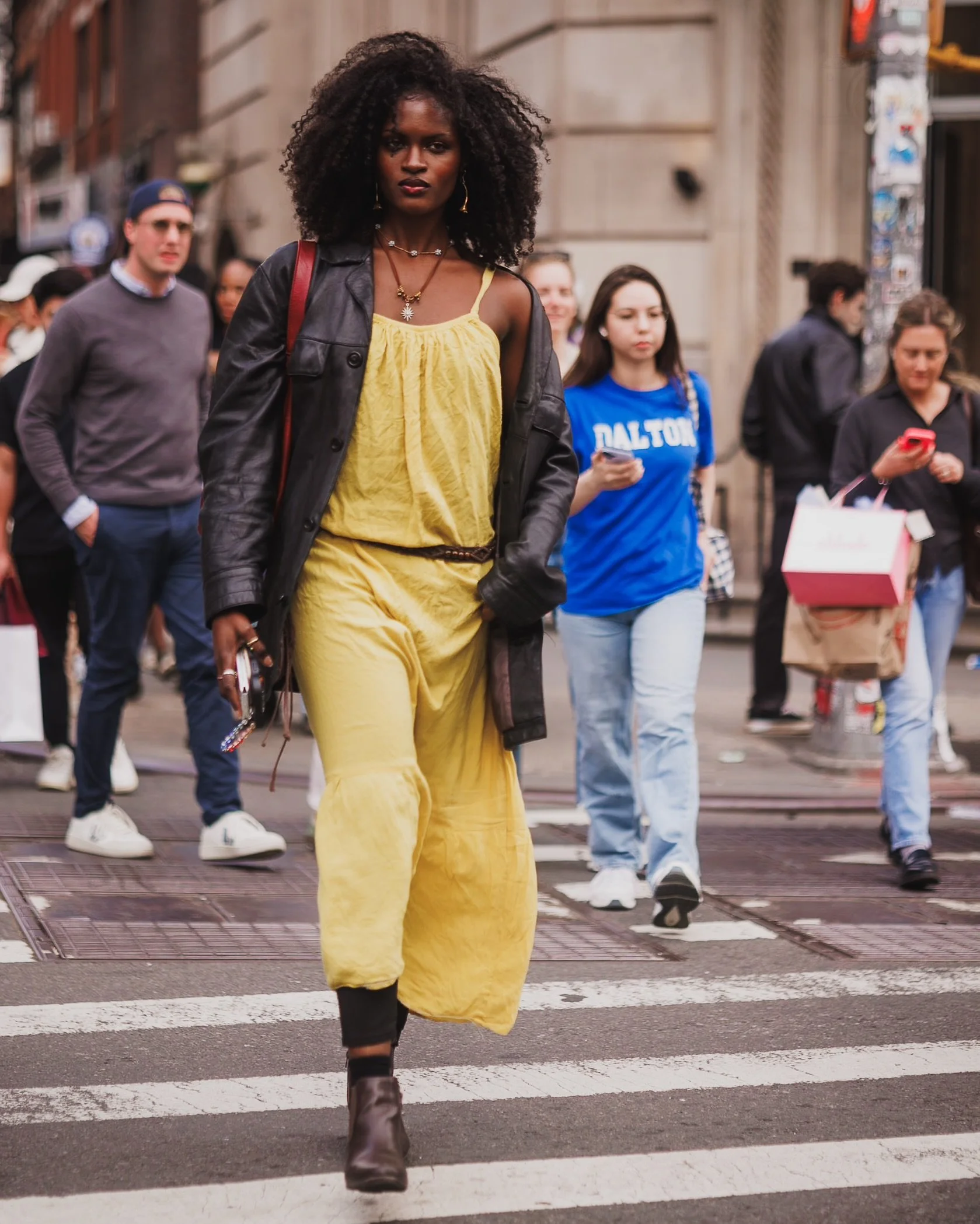 A woman wearing a yellow dress and a black leather jacket crossing a crosswalk in an urban street with a group of people walking behind her.