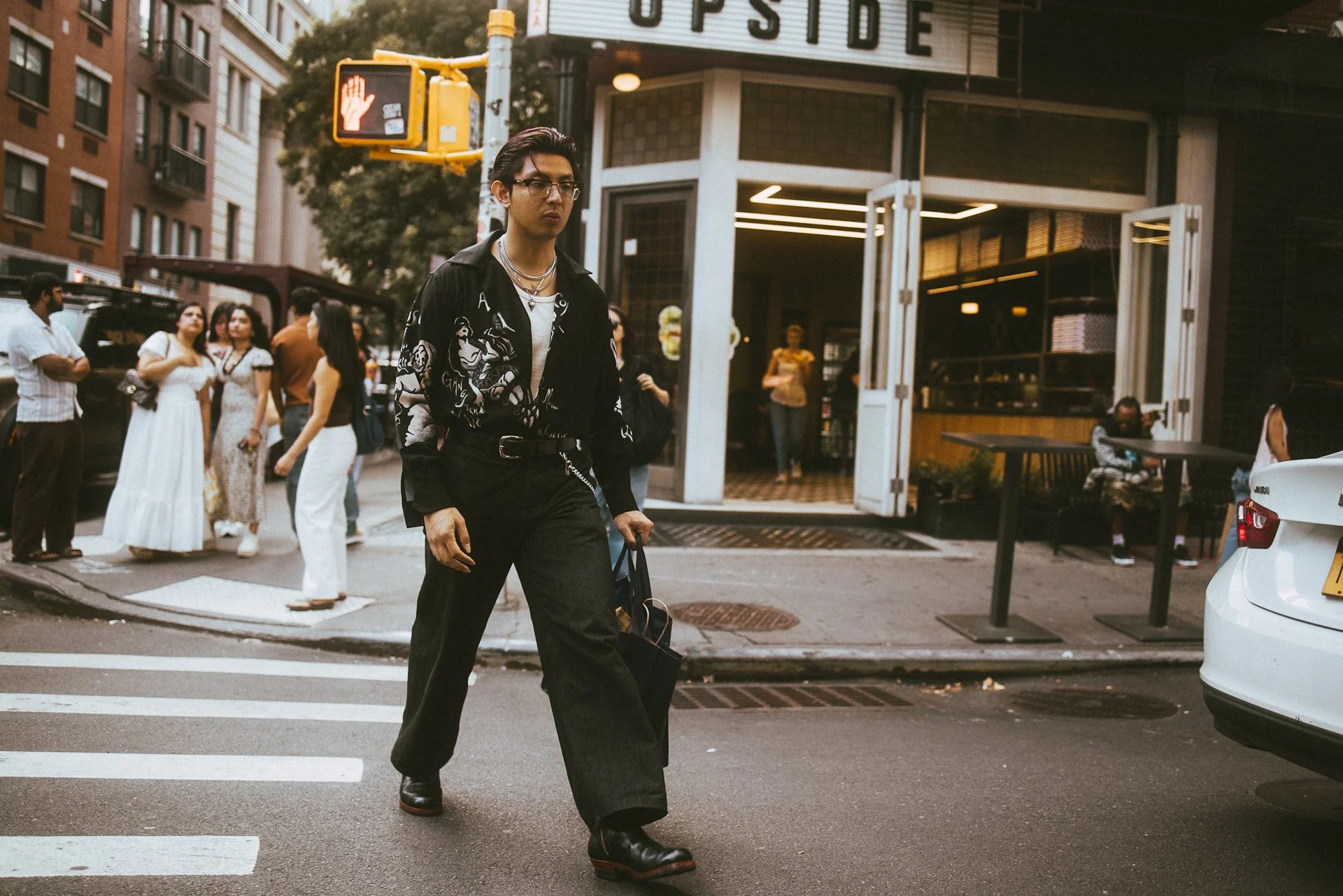 A young man crossing a city street at a crosswalk, wearing black glasses, a black patterned shirt, black pants, and black boots, carrying a black bag in his left hand. In the background, there are several people standing near a building with an open 