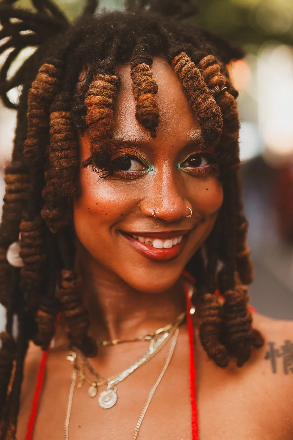 Close-up of a woman with dark skin and styled dreadlocks with light brown tips, smiling and wearing colorful makeup, jewelry, and a red strap across her shoulder.