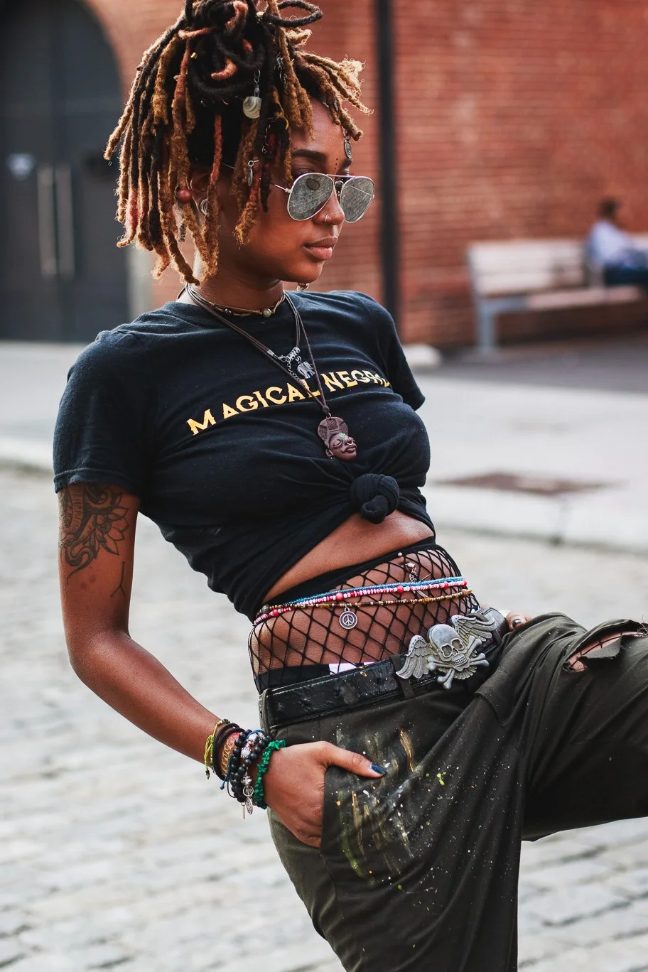 A young woman with dreadlocks wearing sunglasses, a black t-shirt with yellow text, fishnet pants, and multiple colorful bracelets, standing outdoors in an urban setting.