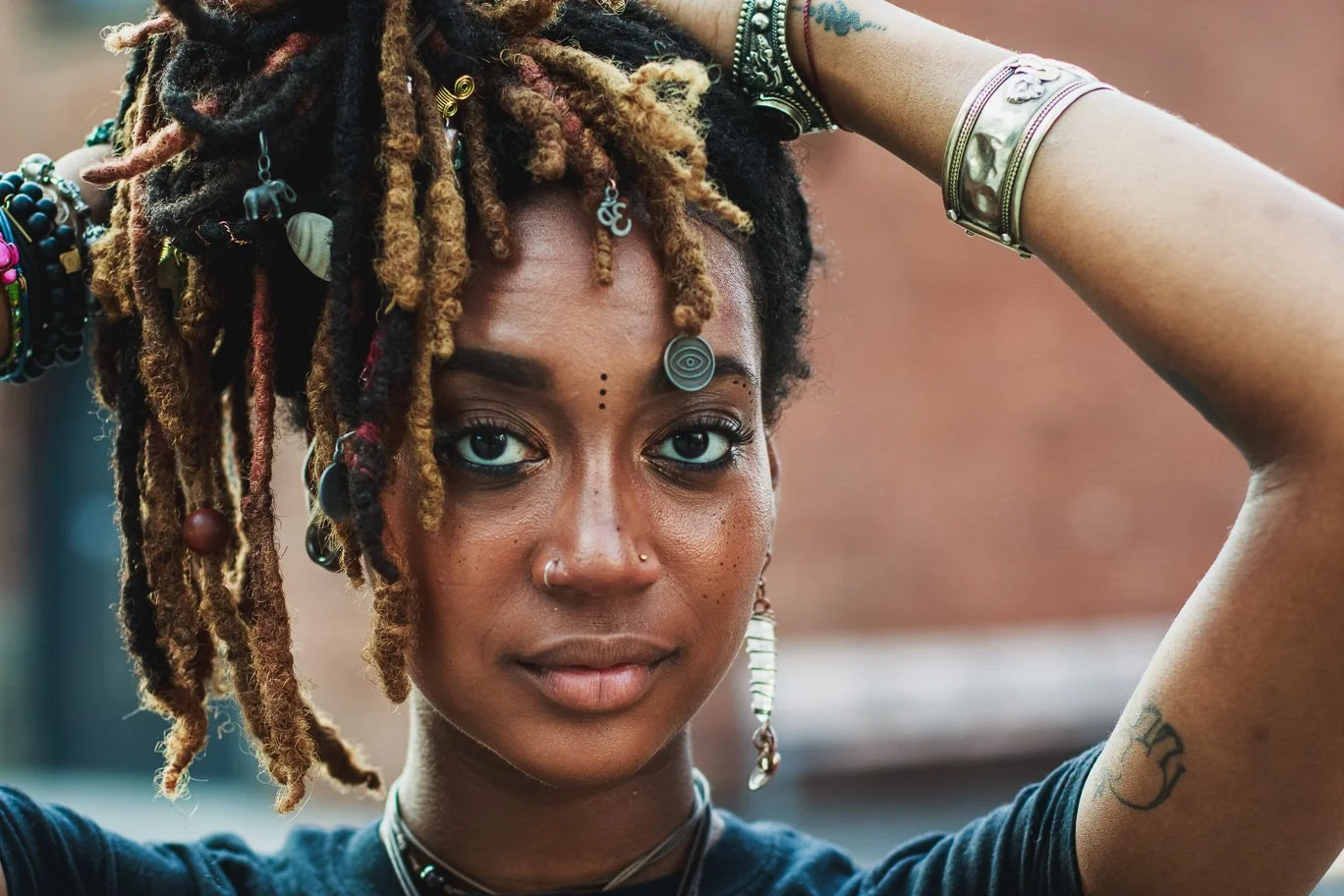 Close-up of a young woman with dreadlocks adorned with jewelry, wearing multiple bracelets and earrings, with a small tattoo on her arm, looking into the camera with a neutral expression.