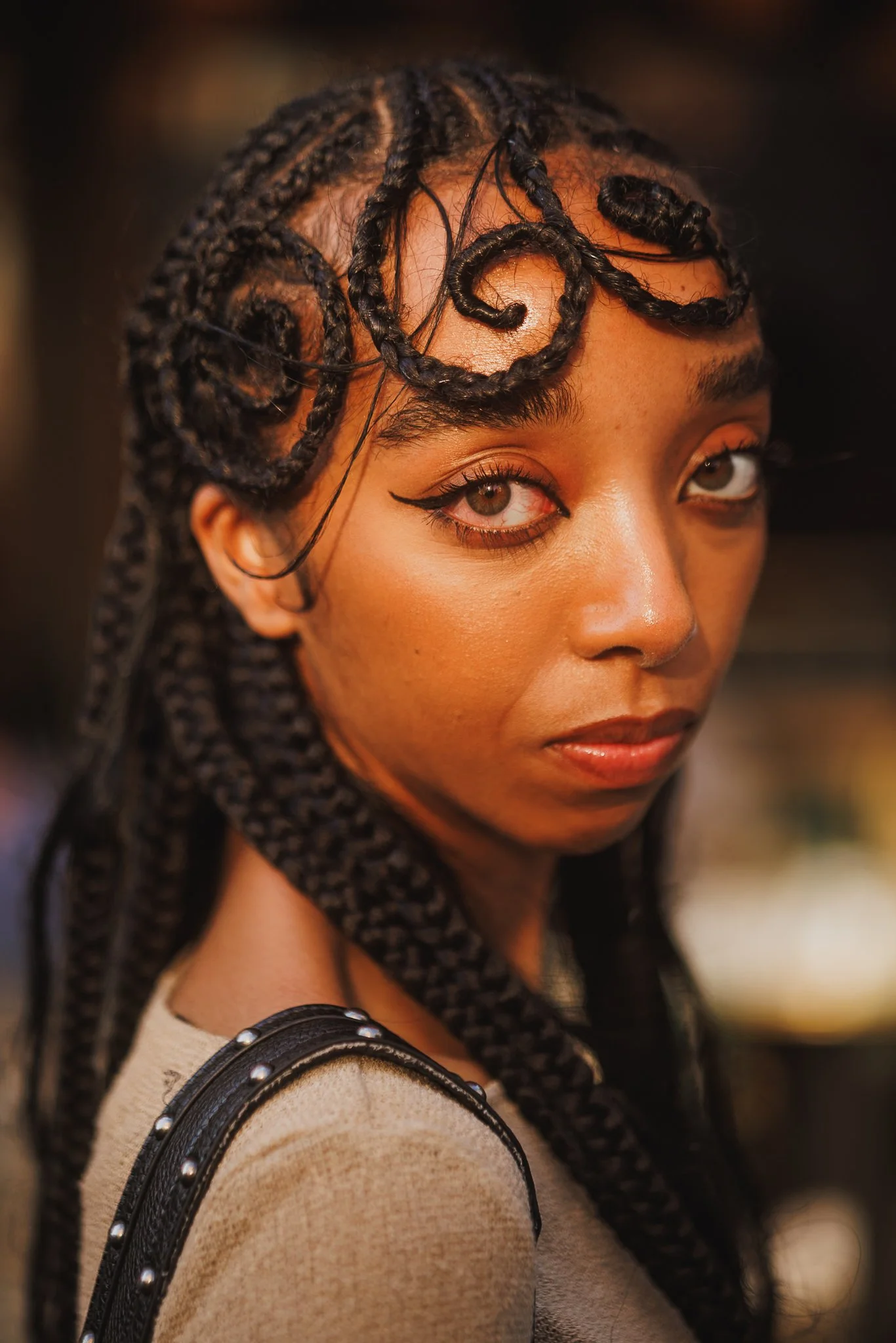 Close-up of a woman with medium skin tone, braided hair, winged eyeliner, and a beige top with black accents and metallic studs on the shoulder.