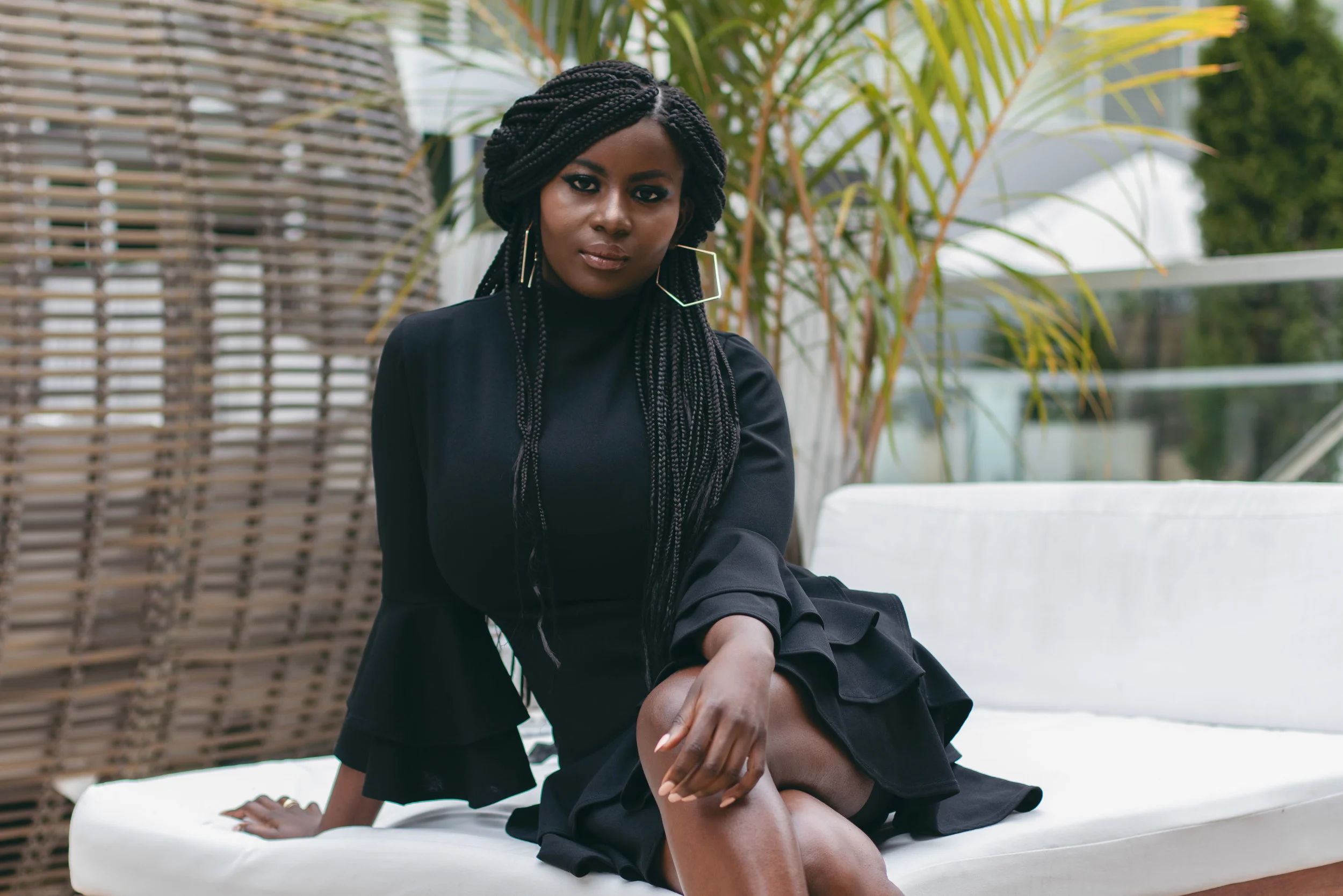 A woman with braided hair and large hoop earrings seated on a white outdoor lounge chair, wearing a black dress with bell sleeves, against a backdrop of green plants and outdoor furniture.
