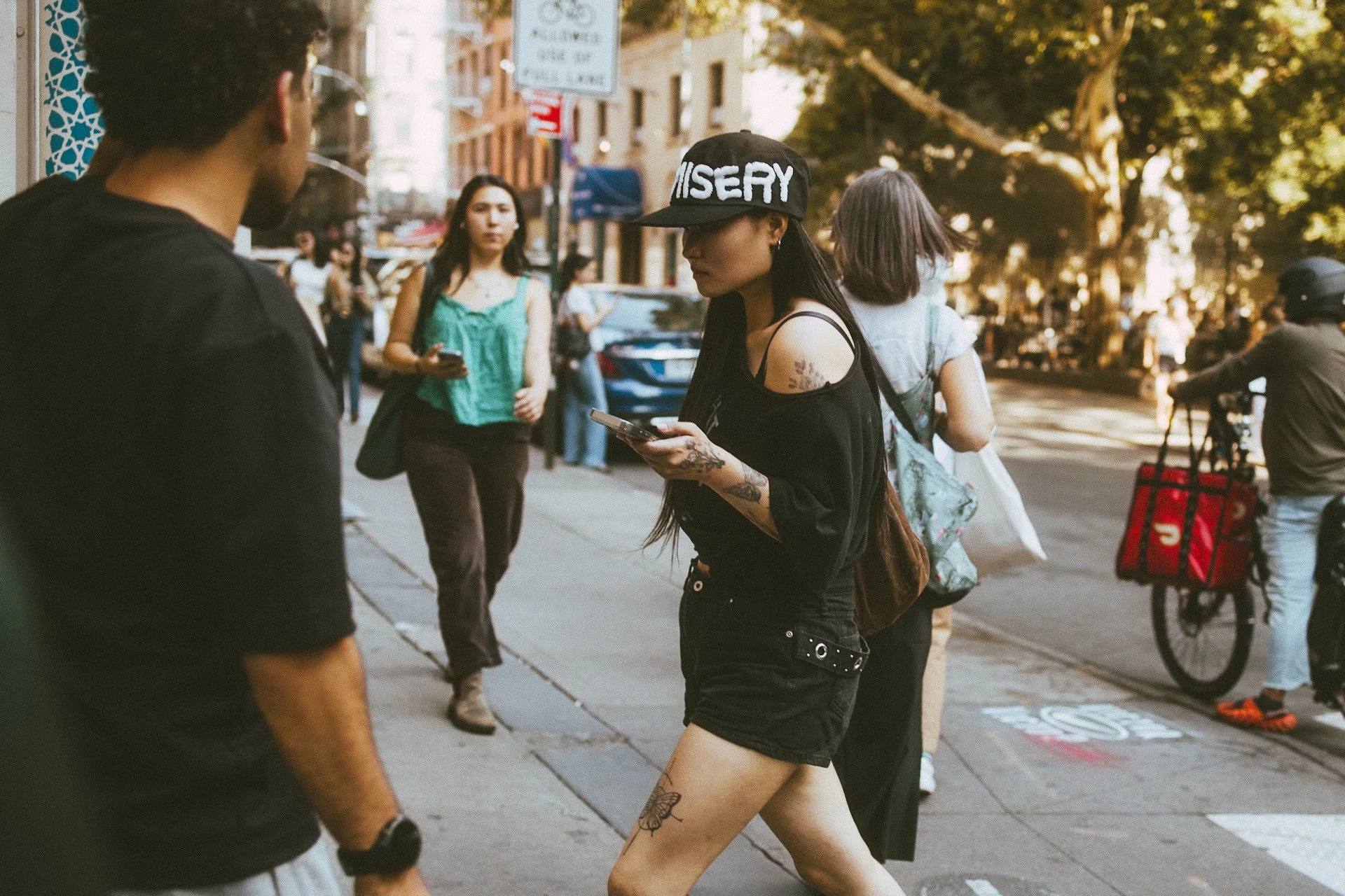 A young woman with tattoos on her arm and leg, wearing a black tank top, black shorts, and a black cap with white lettering, is standing on a city sidewalk looking at her phone. She is surrounded by other pedestrians, some of whom are walking, and th