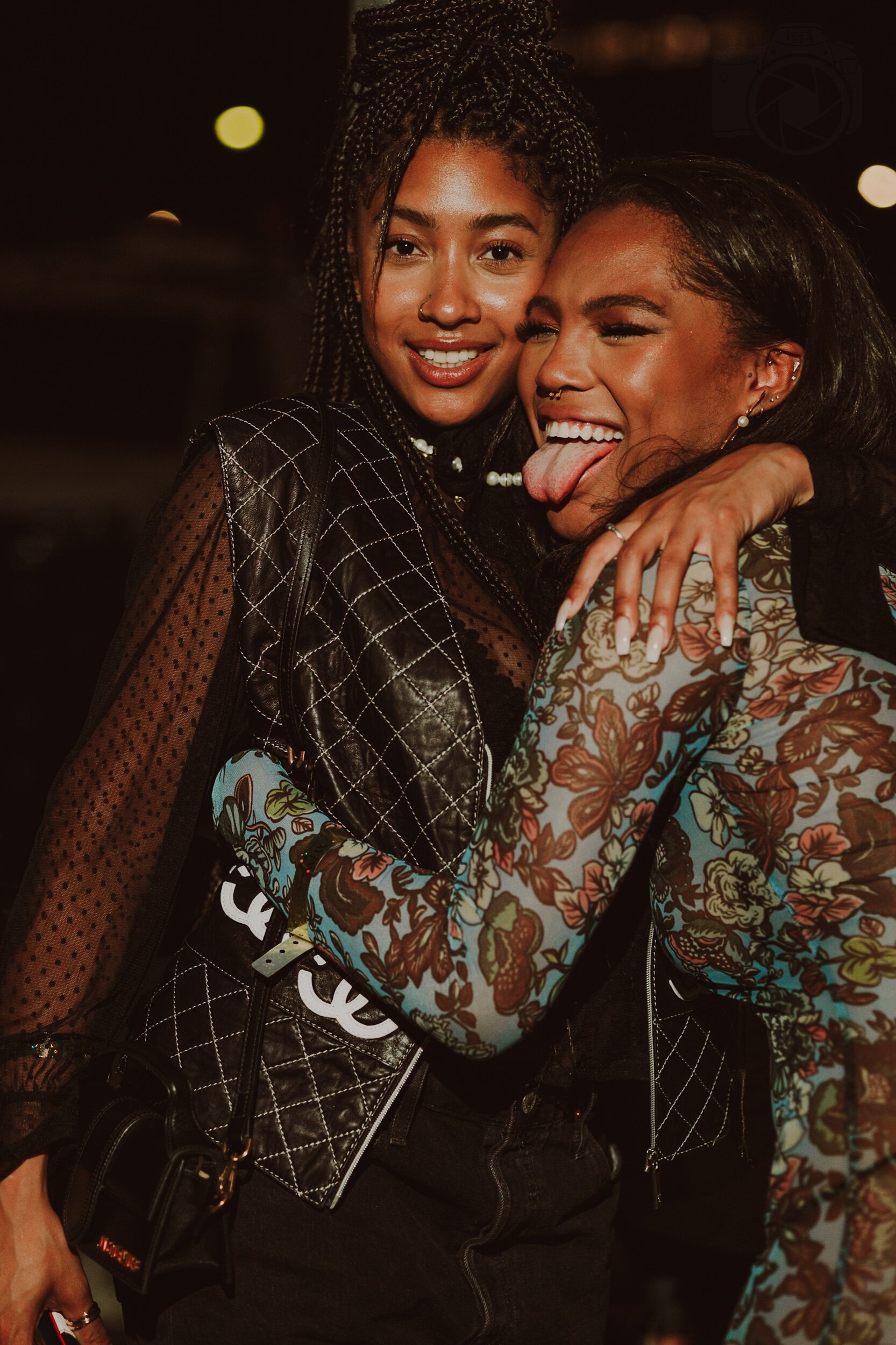 Quen Blackwell with Samira Amalia Ibrahim smiling and hugging each other at night; one sticking her tongue out, wearing colorful floral-patterned jacket, and the other with braided hair, wearing a black quilted leather jacket.