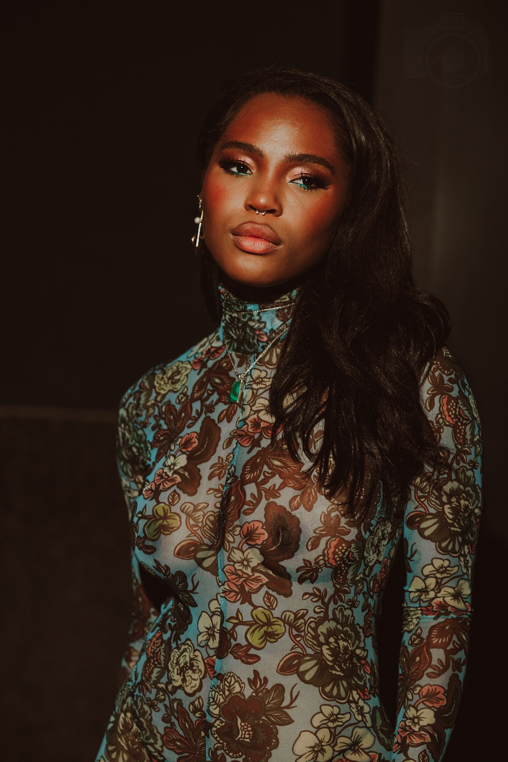 Quen Blackwell with dark skin, long wavy black hair, and makeup, wearing a high-neck floral dress and jewelry, standing against a dark background.