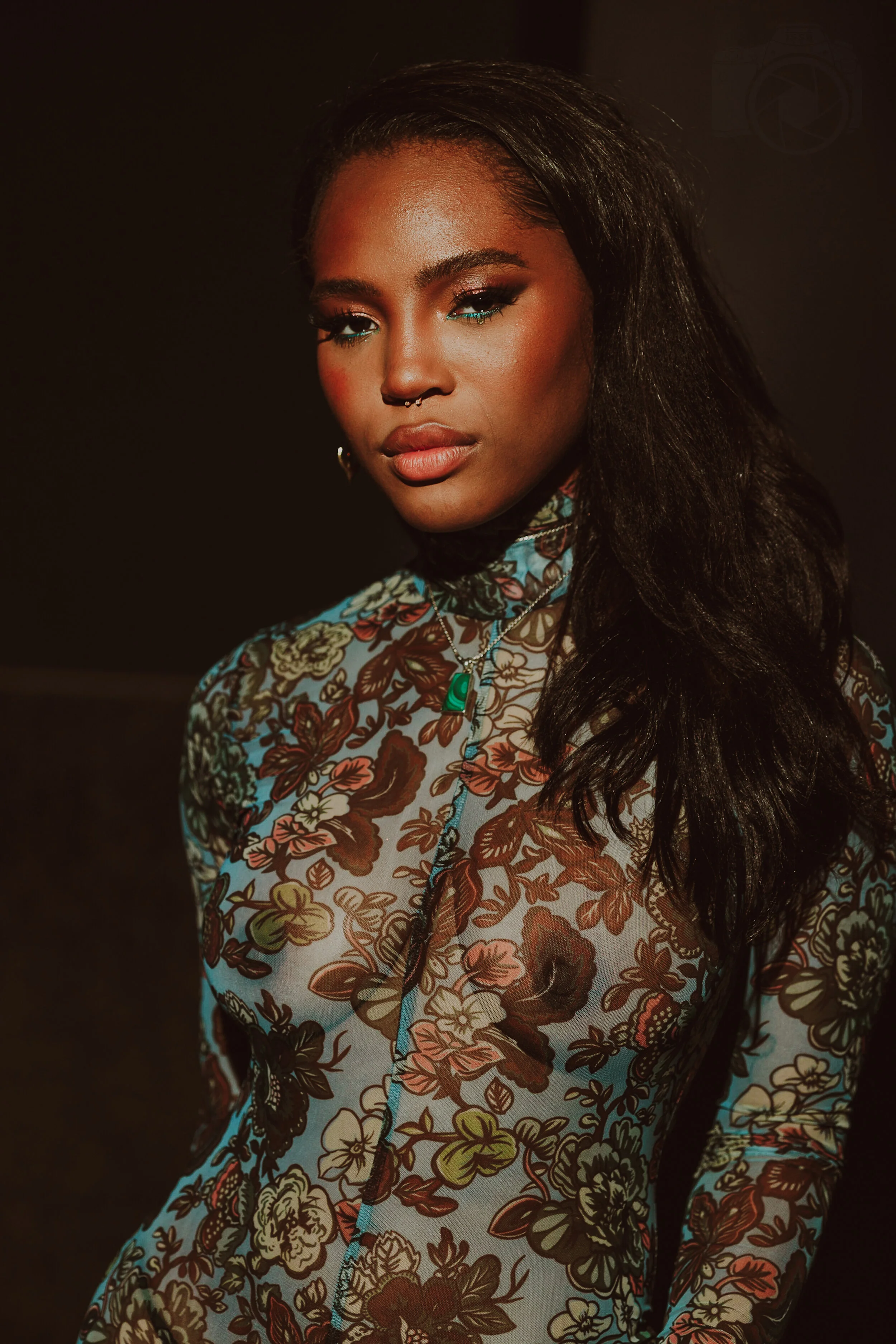 Portrait of Quen Blackwell with dark wavy hair wearing a floral patterned top, standing against a dark background.