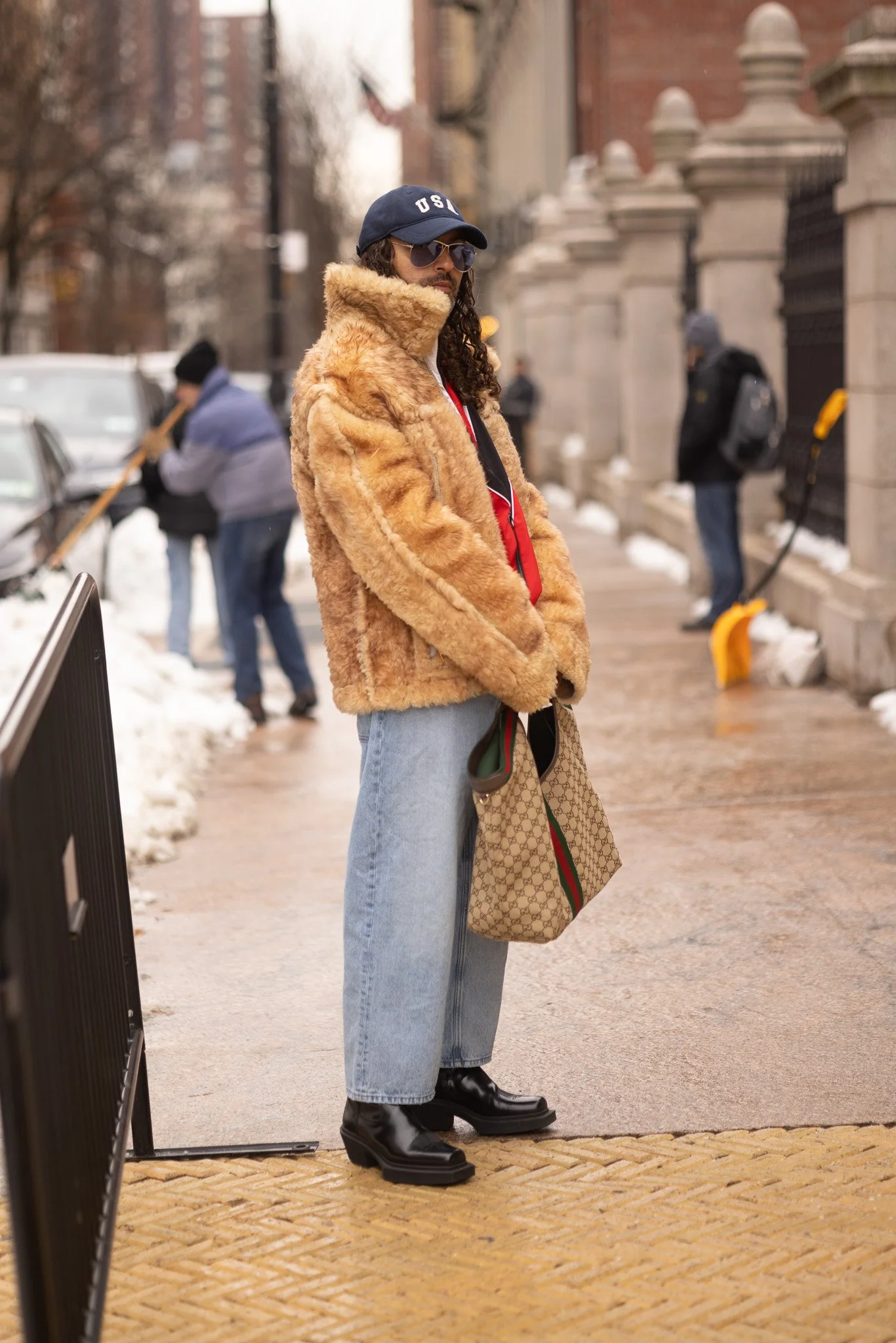 A person standing on a sidewalk wearing a large brown fur coat, light blue jeans, black boots, dark sunglasses, and a blue cap. They hold a beige designer tote bag with a red and green stripe. There are snow piles and other people shoveling snow in t