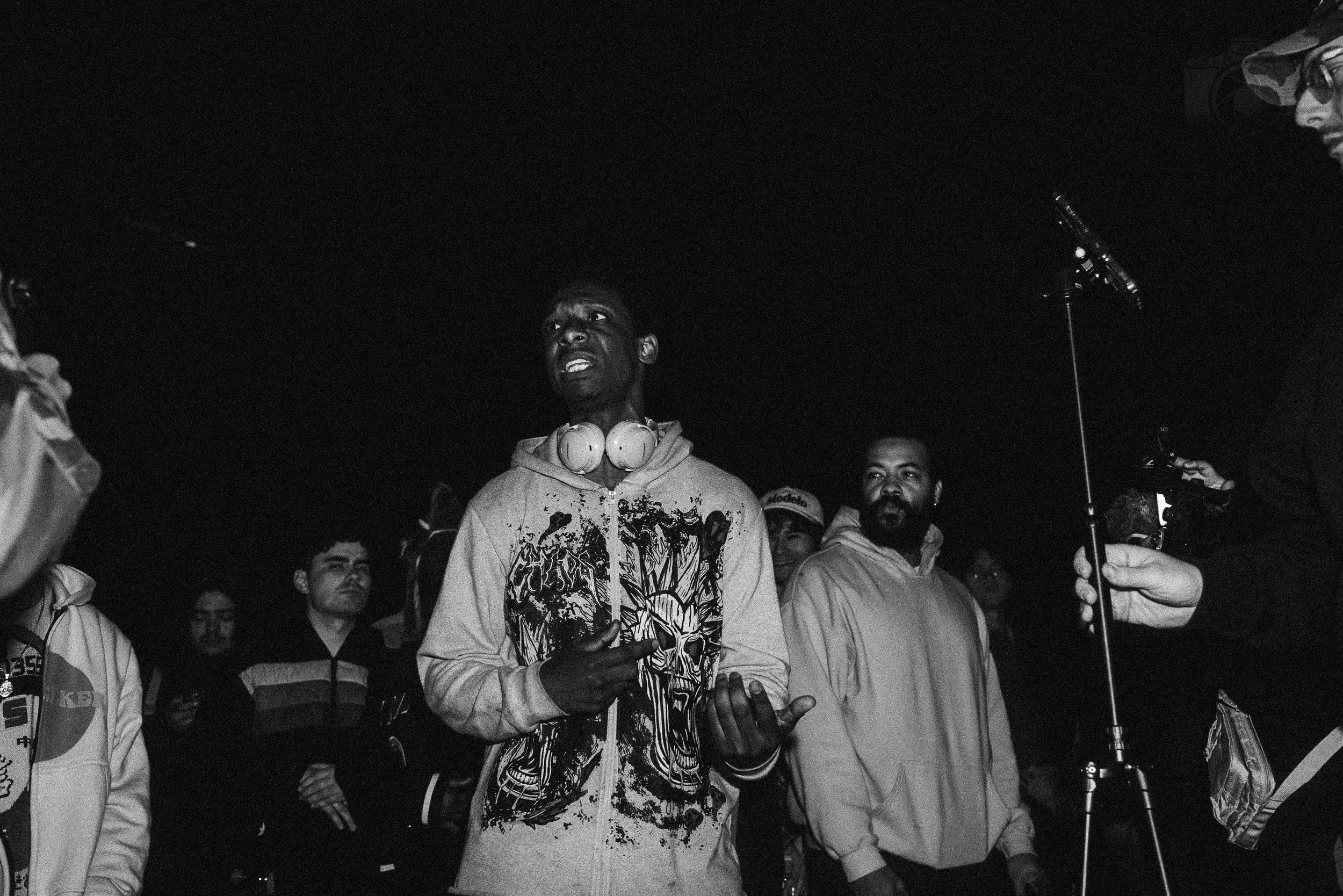 Black and white photo of a young man speaking or singing in front of a crowd, wearing headphones around his neck, with other people around him. It appears to be an outdoor nighttime event.