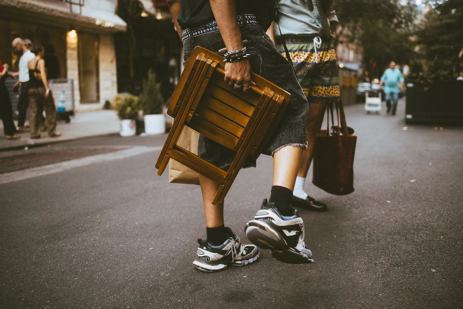 Person riding a skateboard on the street carrying a wooden folding chair, wearing black shorts, a black T-shirt, and sneakers, with other pedestrians in the background.
