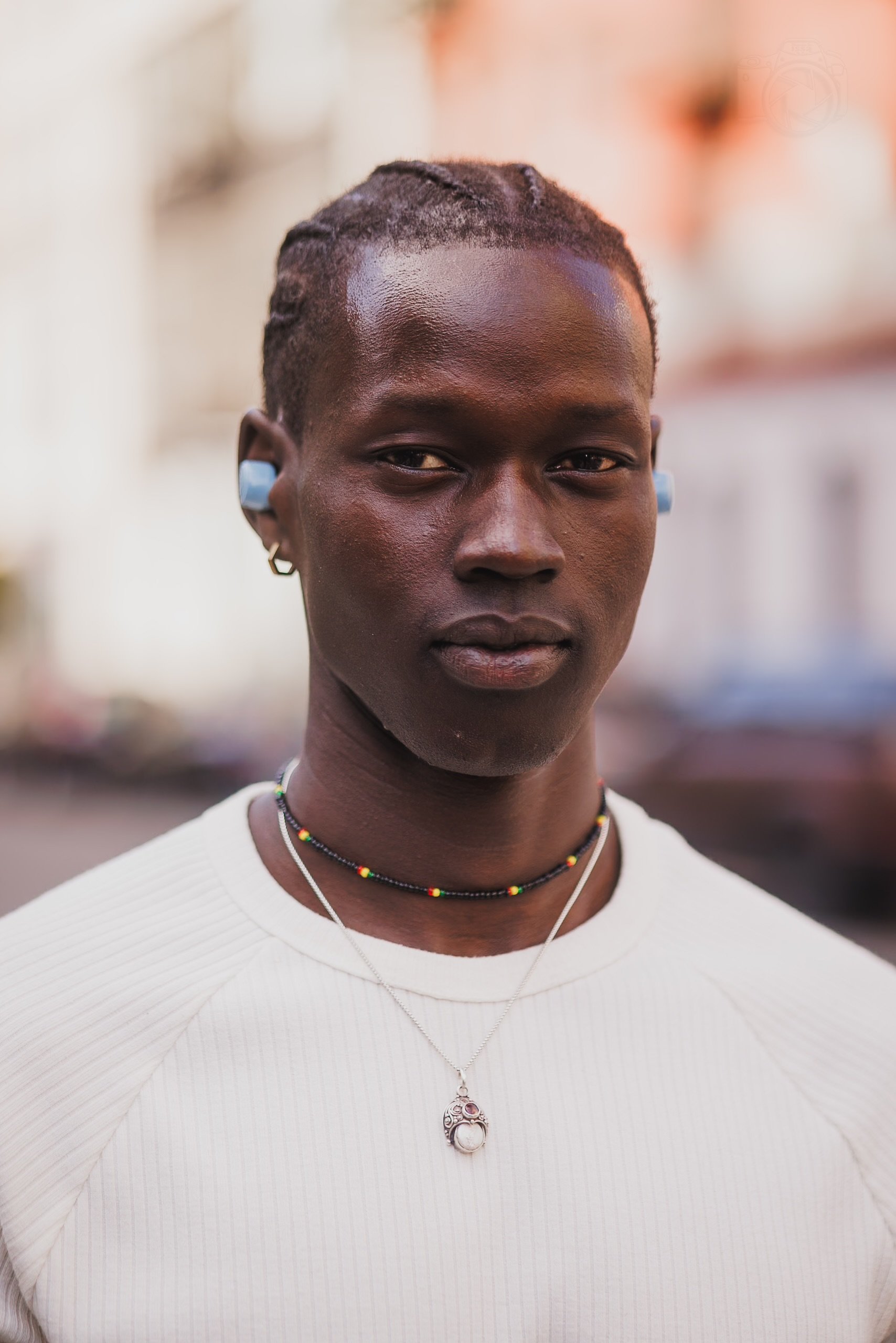 A young Black woman with short, braided hair, wearing a white top, colorful beaded necklaces, a silver pendant necklace, a small hoop earring, and wireless earbuds, standing outdoors with a blurred background.