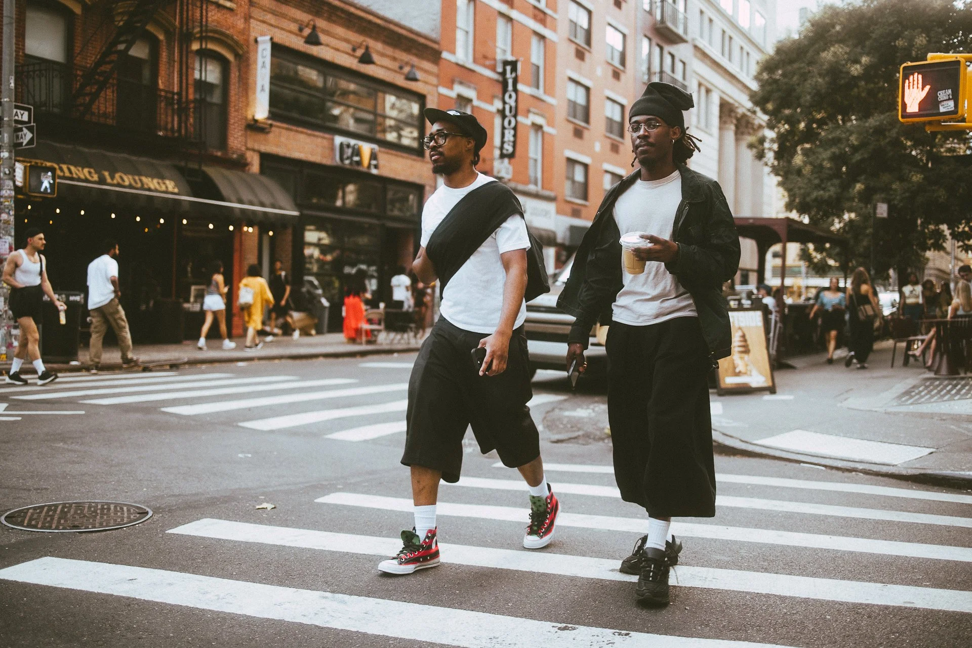 Two young men crossing a city street during daytime, walking on a crosswalk. One is wearing a black jacket, white shirt, black pants, and black shoes, holding a cup of coffee. The other is dressed in a white T-shirt, black shorts, and colorful sneake