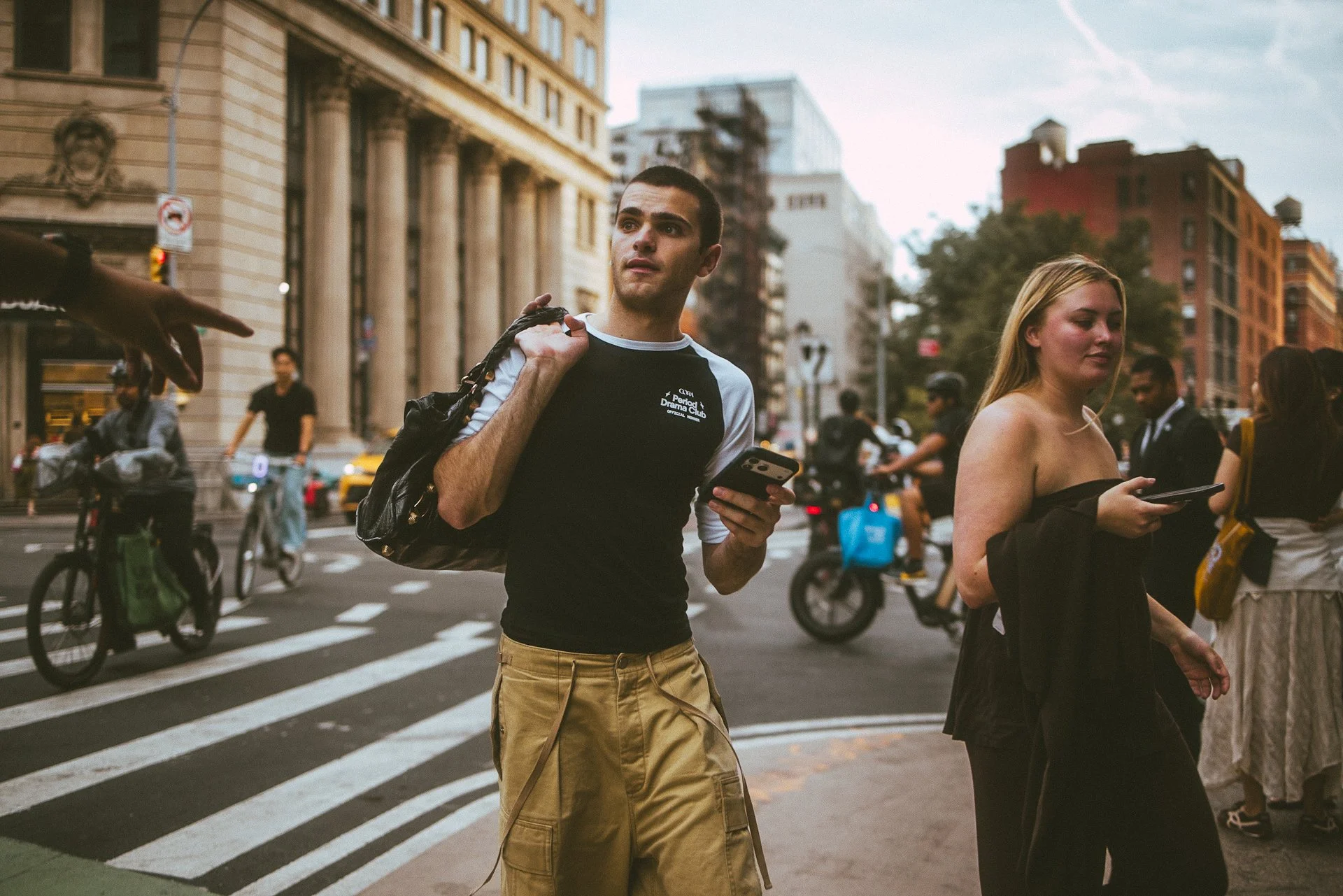 Young man and woman waiting at a crosswalk on a busy city street with buildings, cyclists, and pedestrians around.