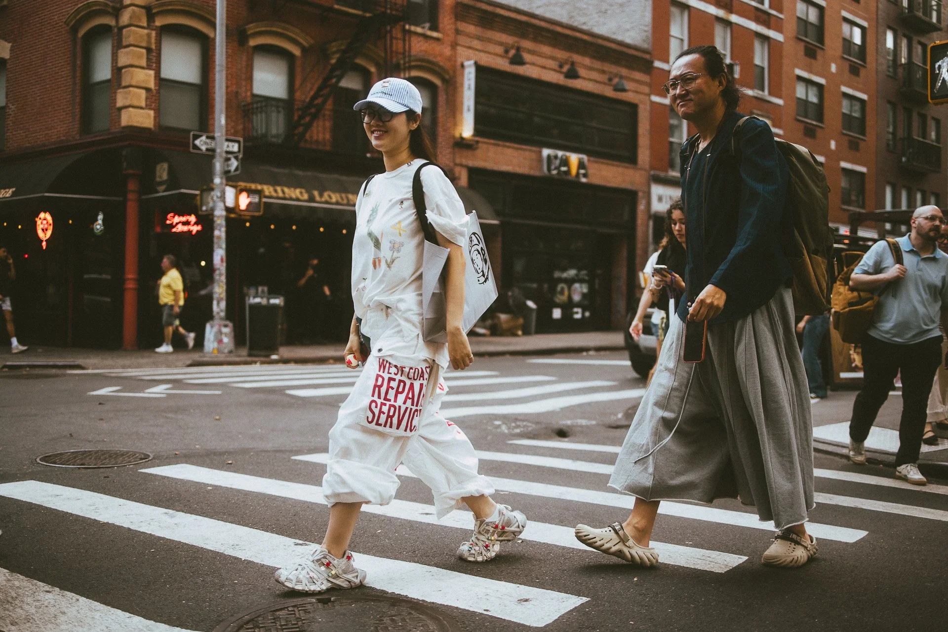 People crossing a city street with brick building in the background, including a woman in white pants and a white shirt with colorful designs, and another woman in gray skirt and dark top, with other pedestrians and storefronts in the scene.