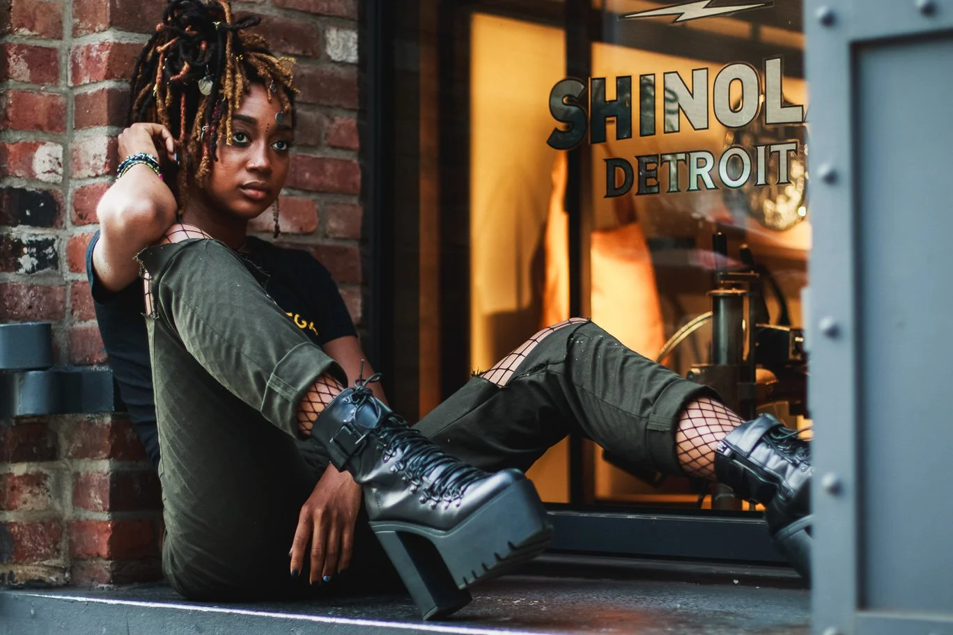 A young woman with dreadlocks and dark clothing sitting against a brick wall outside a shop with a glass window that has the words 'Shinol Detroit' on it.