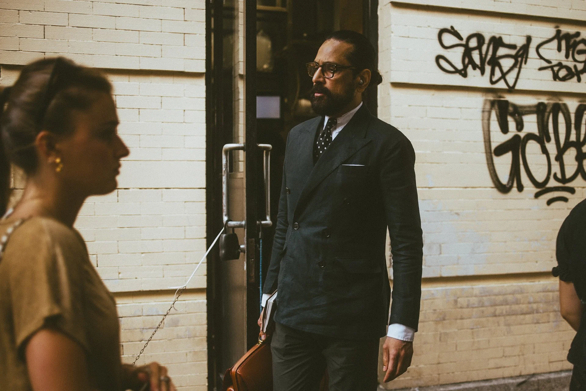 A man with glasses, a beard, and slicked-back hair, dressed in a dark suit with a polka-dot tie, standing on a city street near a brick wall with graffiti.