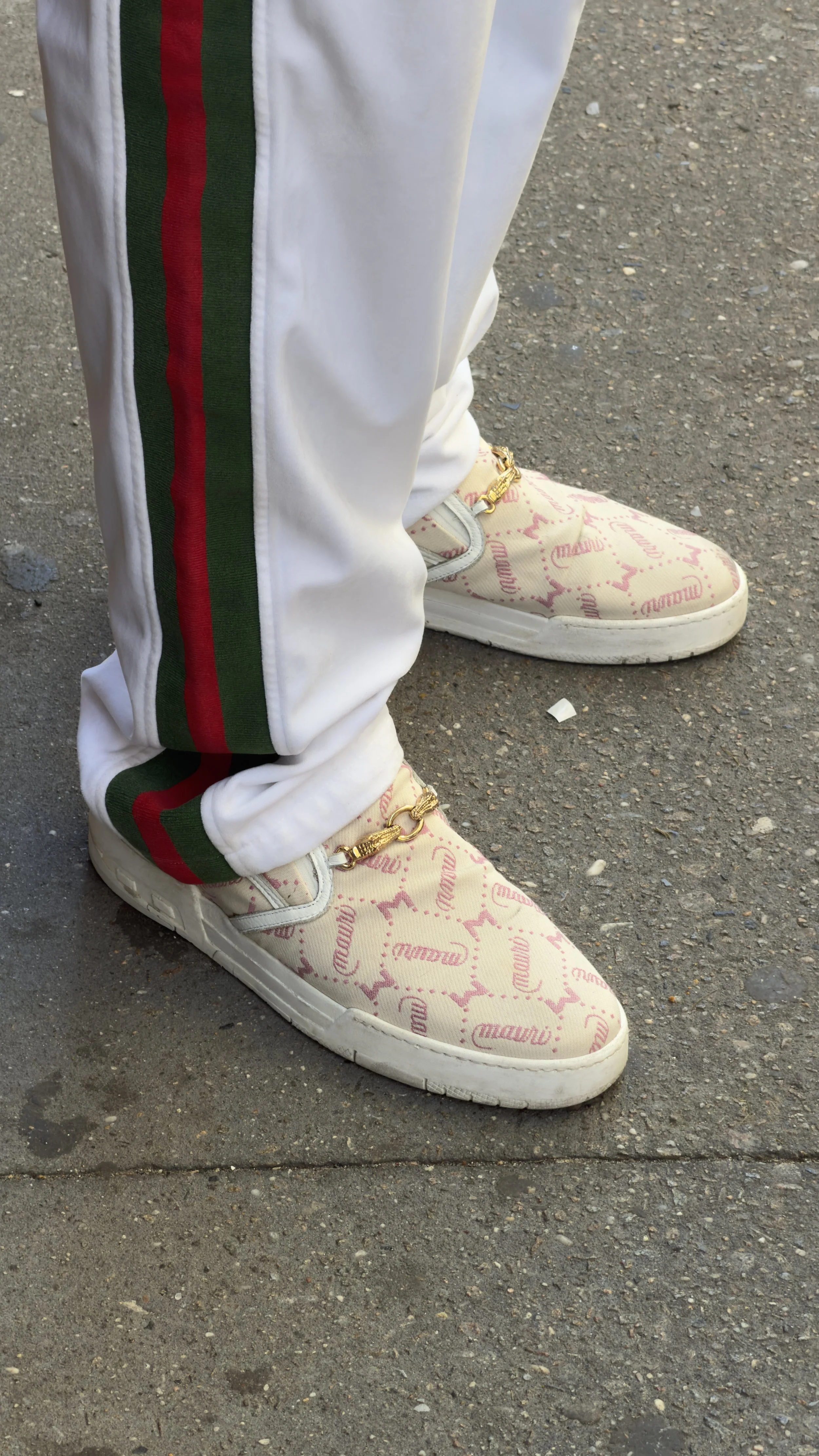 Close-up of person wearing white pants with red and green stripe, golden chain on white sneakers featuring pink logo text, standing on asphalt ground.