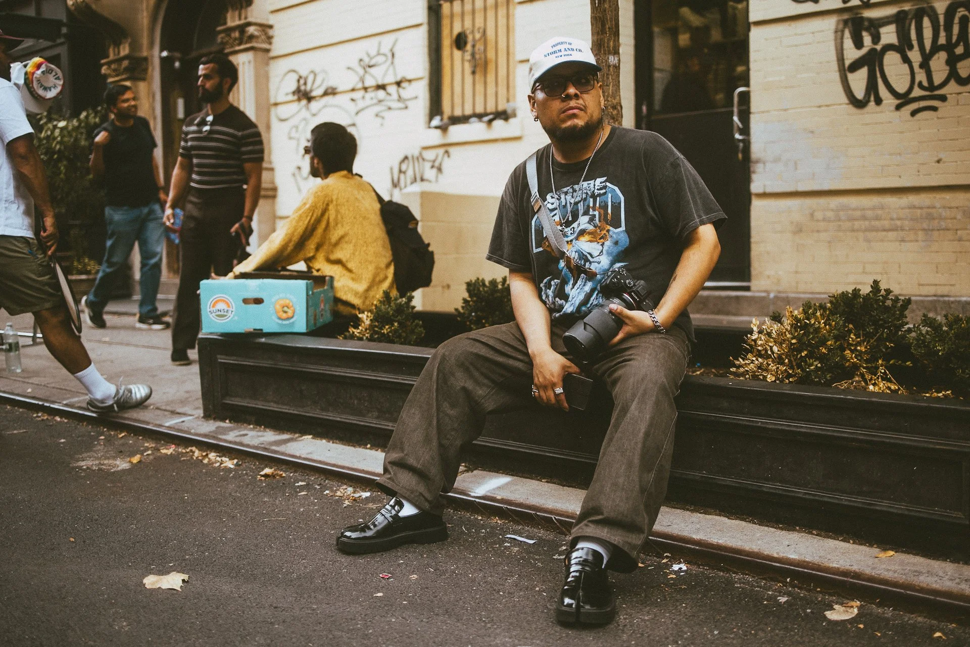 A man sitting on a black bench on a city street, holding a camera. He wears a black graphic t-shirt, brown pants, shiny black shoes, sunglasses, a baseball cap, and jewelry. In the background, there are people walking and talking near a building with