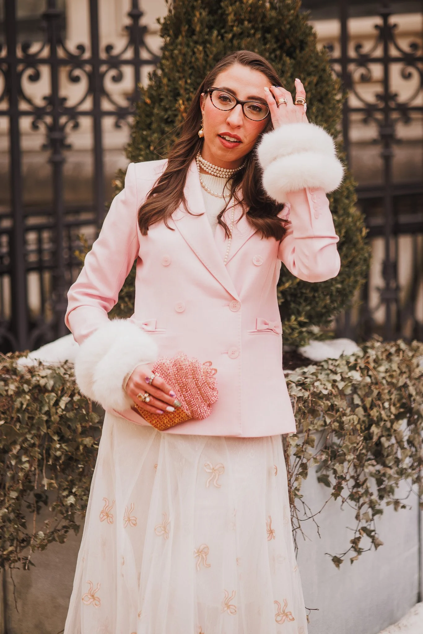Caroline Vazzana dressed in a pink blazer with white fur cuffs, a cream tulle skirt with bows, and layered pearls, holding a pink beaded clutch, standing outdoors in front of a black metal fence and bushes.