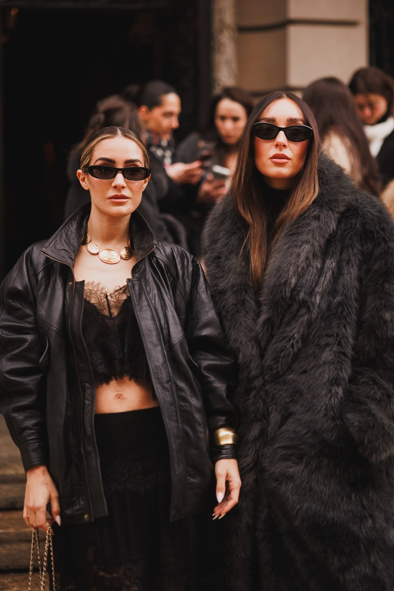 Two women standing outdoors, wearing dark, fashionable clothing and sunglasses, with a group of people in the background.
