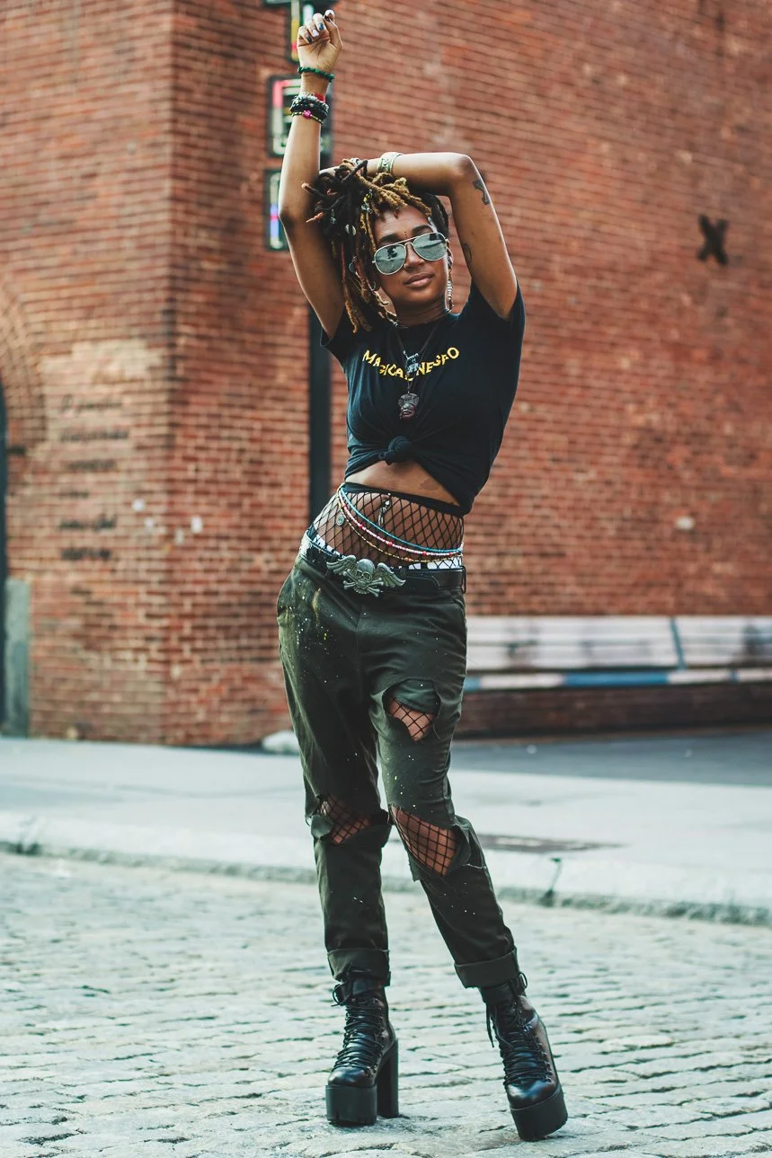 A young woman with dreadlocks and sunglasses poses with arms raised above her head in front of a brick wall. She is wearing a black knotted T-shirt, fishnet pants over ripped jeans, and high-heeled boots, accessorized with jewelry and bracelets.