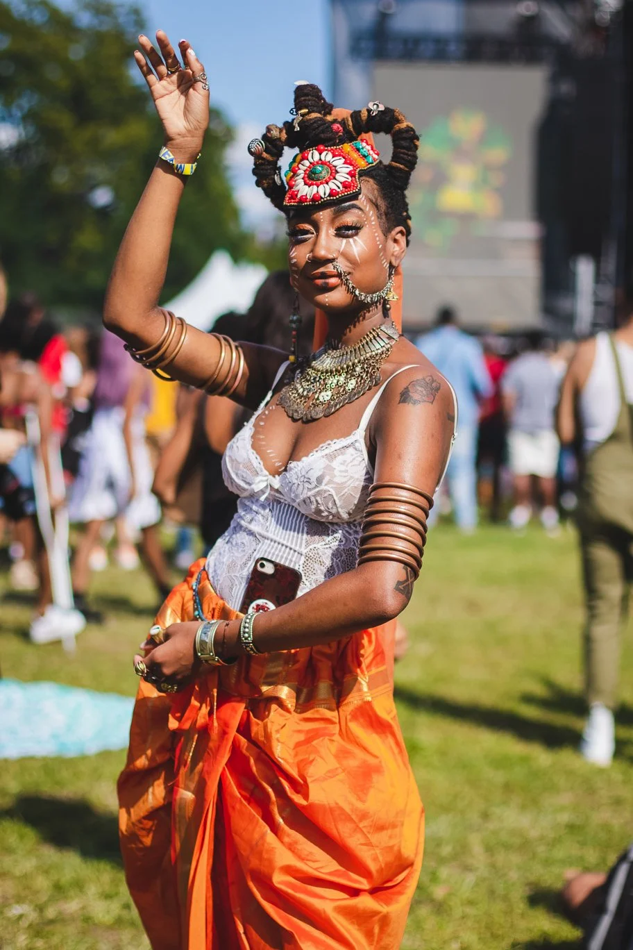 A young woman with elaborate hair and jewelry dancing outdoors at a cultural festival, wearing a white lace top and orange skirt.
