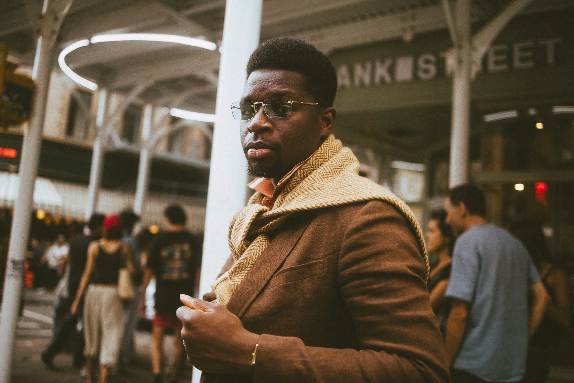 A man with dark skin, wearing sunglasses and a brown blazer with a beige patterned scarf, is standing in a busy indoor public space, possibly a train station or airport, with people walking in the background and illuminated curved ceiling structures.