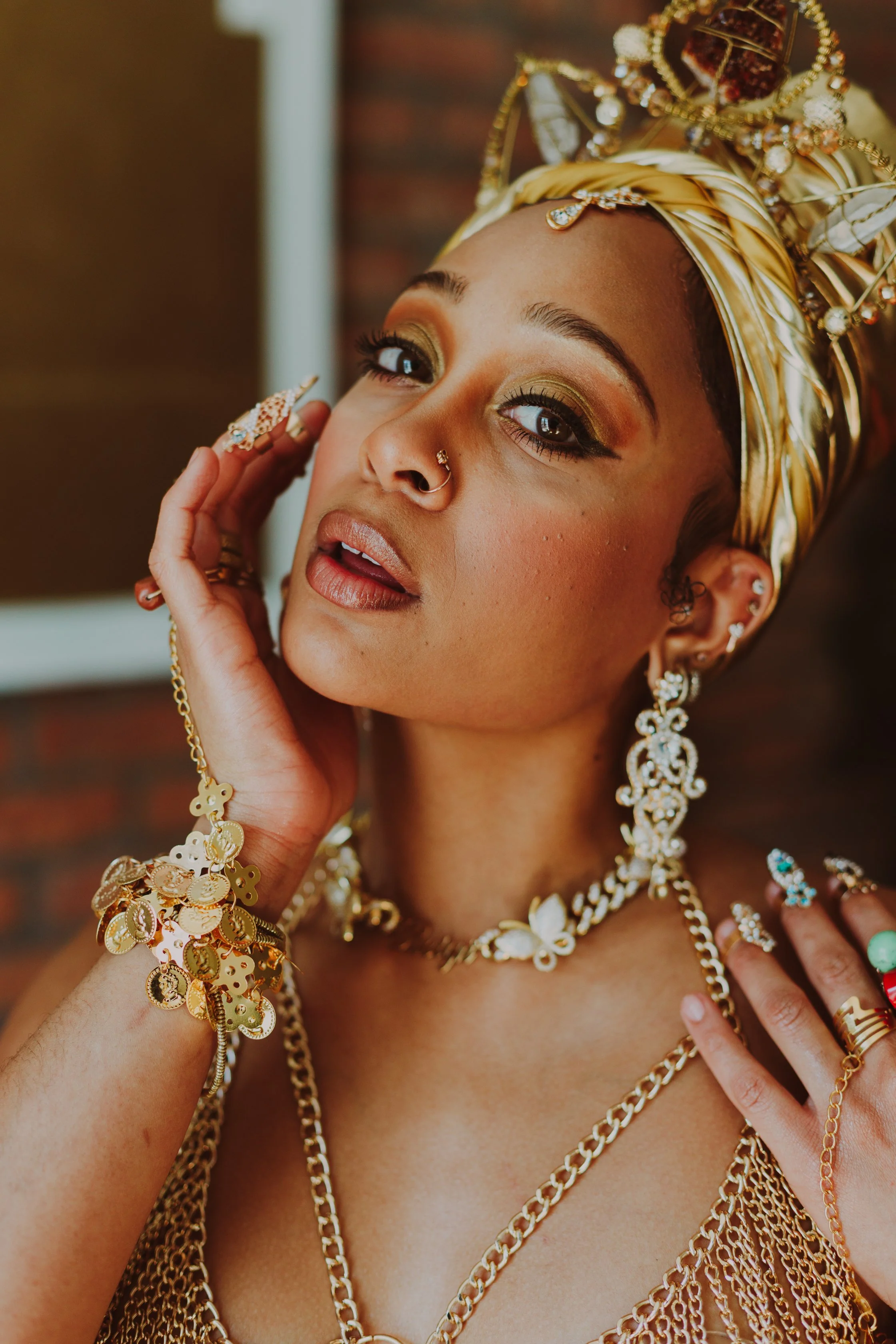 A woman with gold jewelry, including necklaces, earrings, rings, and a head wrap with gold and white accents, posing with her hand near her face.