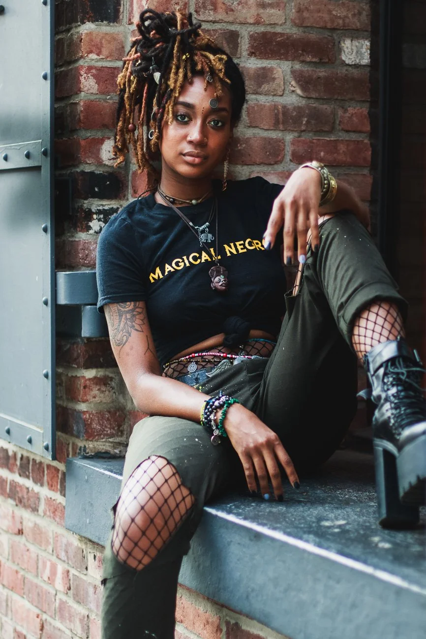 A young woman with dreadlocks sitting against a brick wall, wearing a black t-shirt with yellow text, green ripped pants paired with fishnet stockings and black lace-up boots, accessorized with layered necklaces, bracelets, and earrings.