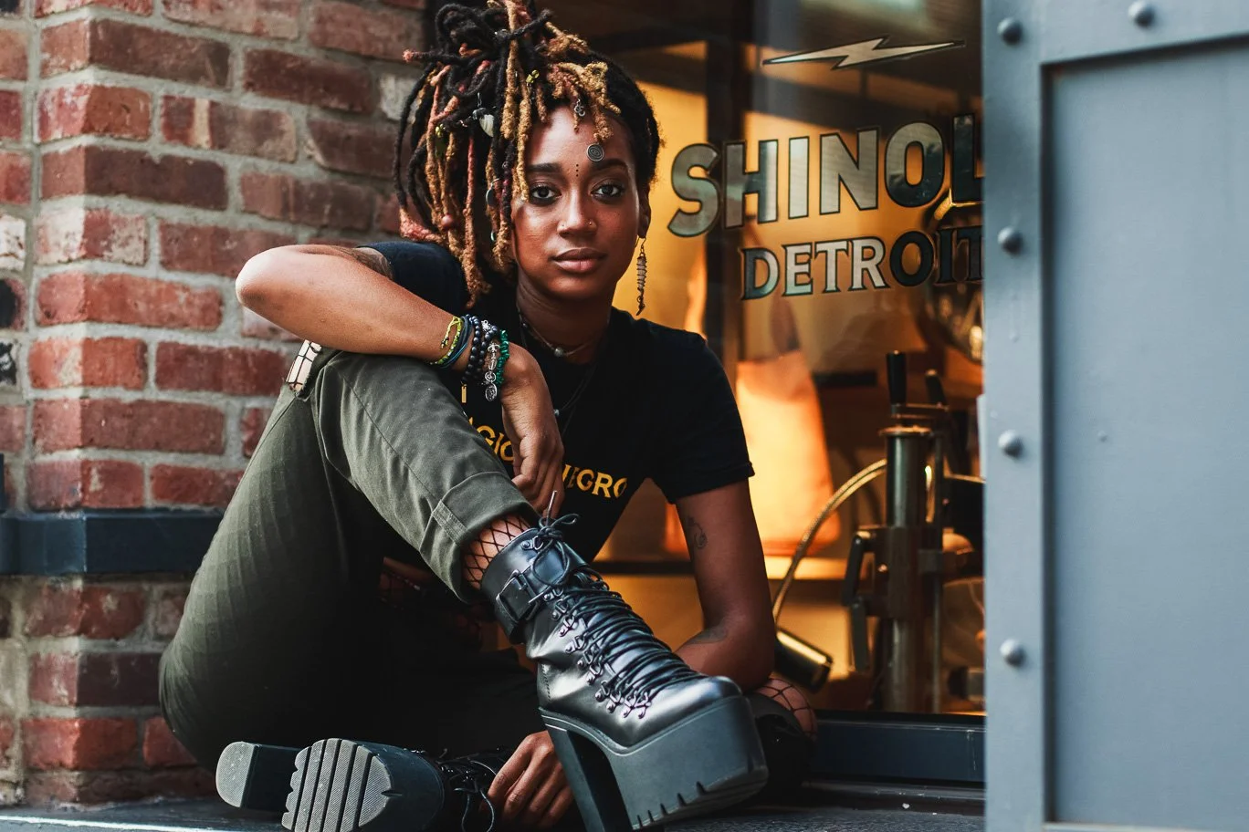 A young woman with dreadlocks and piercings sitting outside a shop named 'SHINOL DETROIT', wearing black clothing, boots, and colorful jewelry, with a brick wall background.