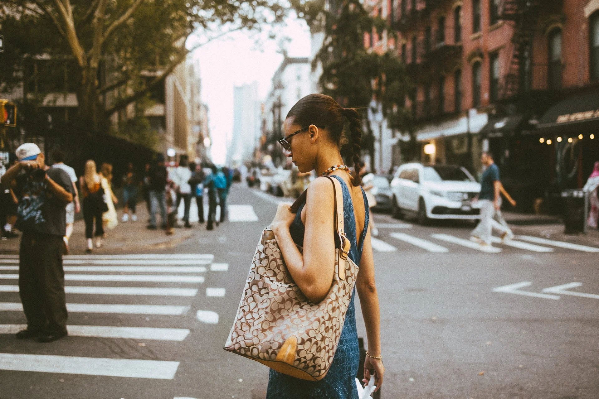 A woman with dark hair in a braid, wearing sunglasses, a blue dress, and a necklace, is crossing a city street with a large patterned bag over her shoulder. She is surrounded by pedestrians and parked cars with buildings and trees in the background.