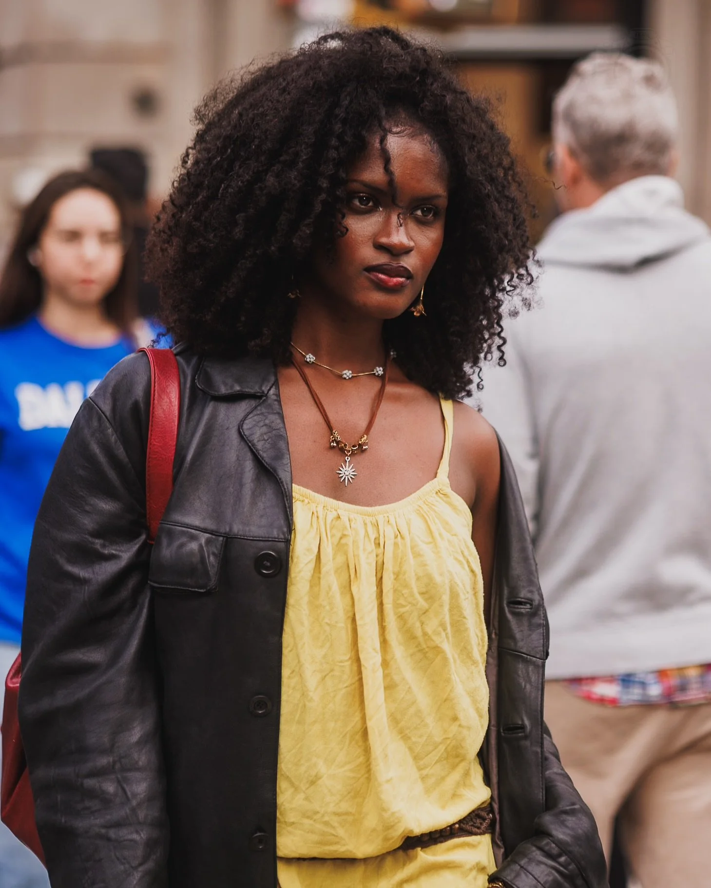 A Black woman with curly hair, wearing a yellow dress, a black leather jacket, and gold jewelry, at a crowded indoor setting.