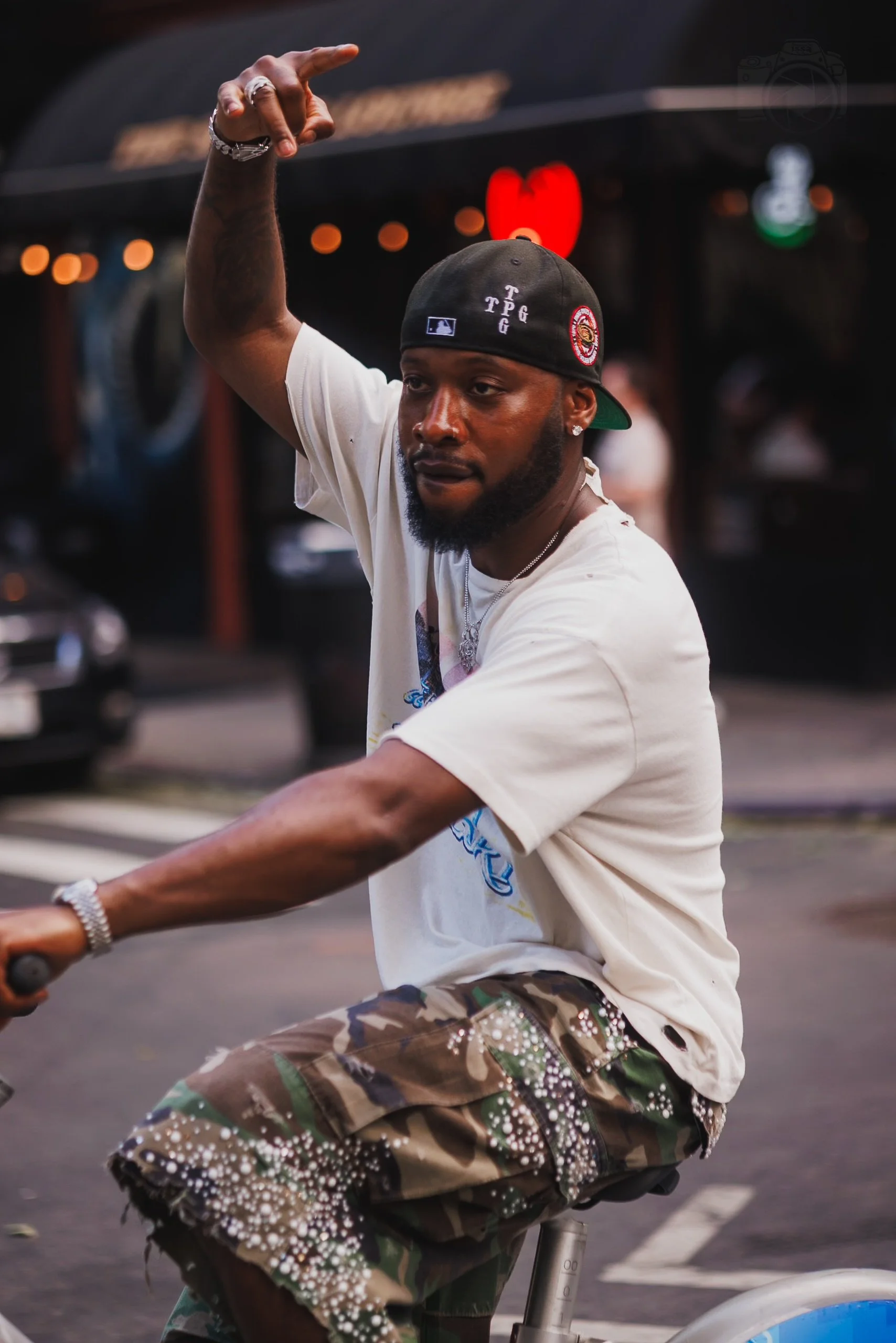 A man riding a bicycle, wearing a black baseball cap turned backwards, white t-shirt, and camouflage shorts with white embellishments, is pointing with his right hand. The background shows a blurry parking lot and a store with a red heart-shaped sign