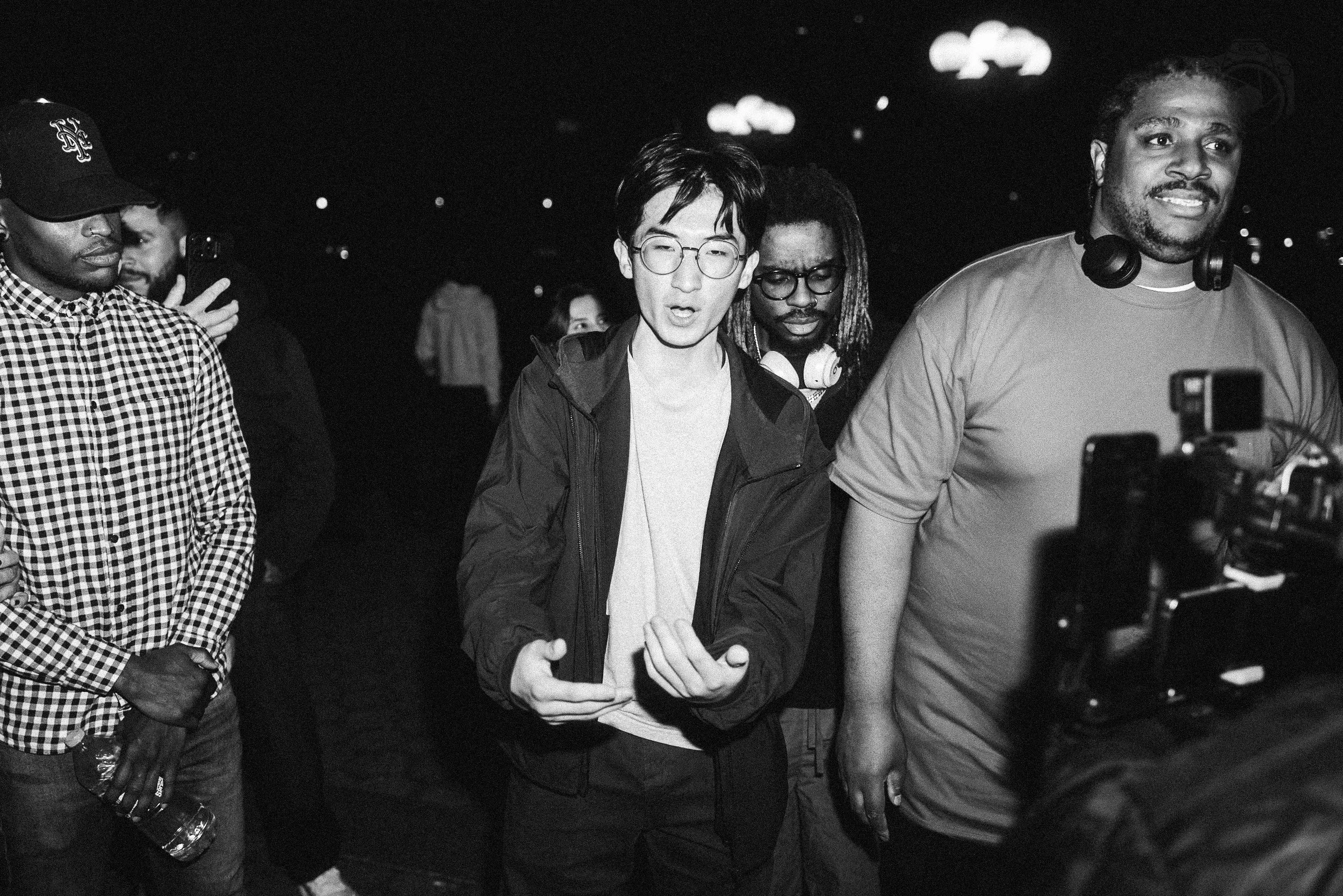 A group of five diverse people, including a woman with glasses, a woman with dreadlocks, and a man with headphones around his neck, standing together and looking at the same point, appear to be at a social event or gathering in a dimly lit environmen