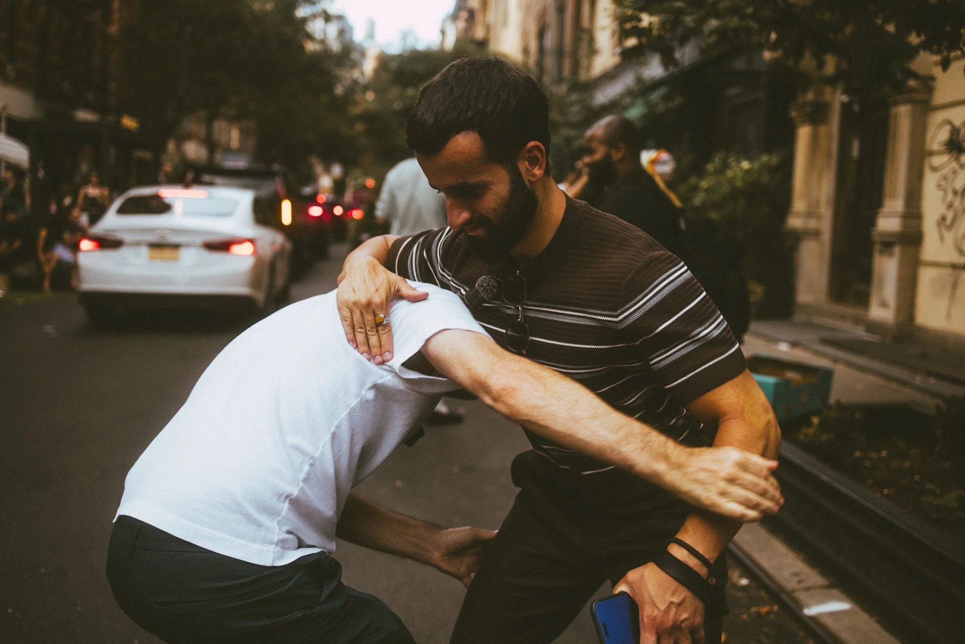 Two men are involved in a physical altercation on a city street, with one man grabbing the other around the neck and shoulder. The scene appears tense, with other people and cars visible in the background.