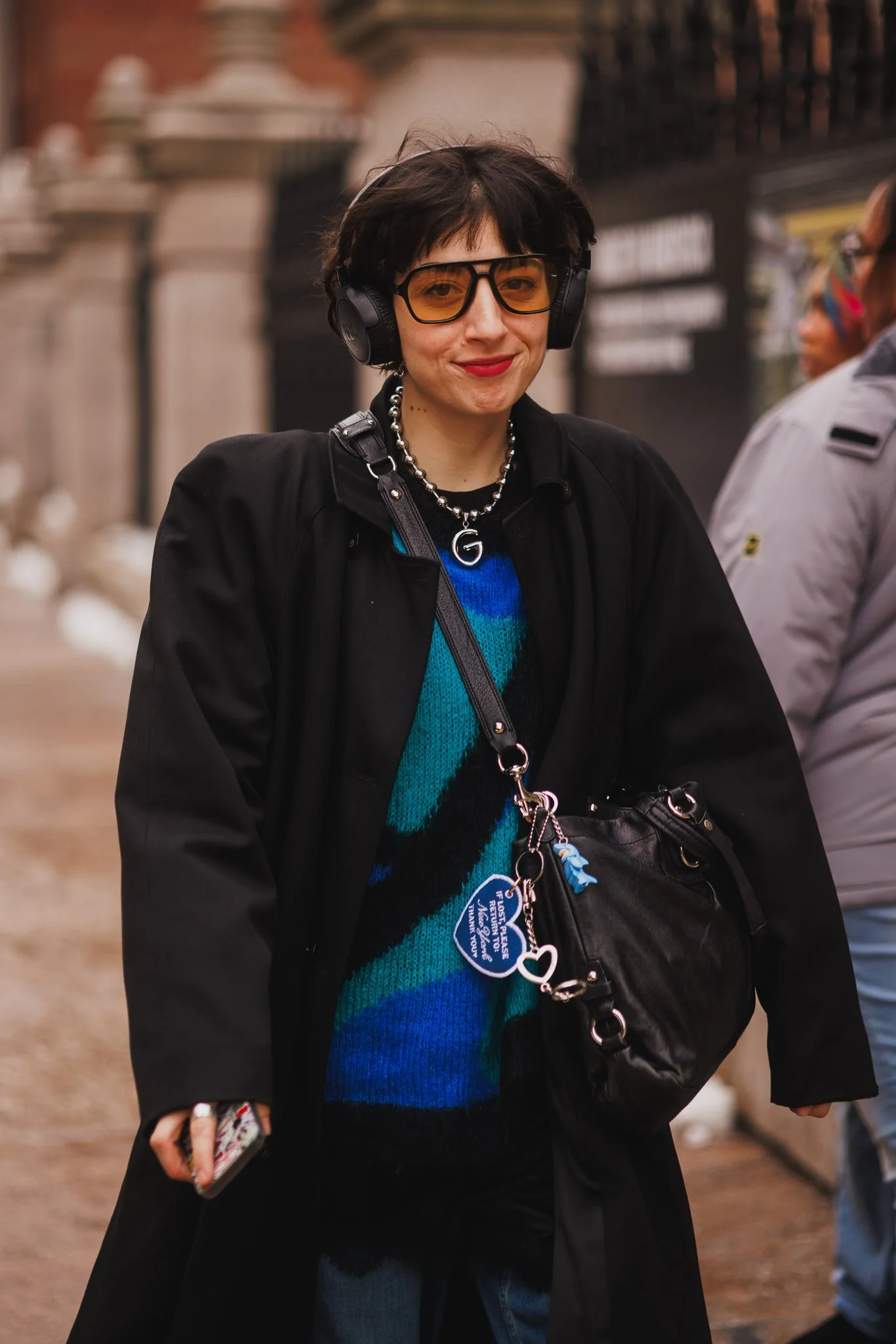 A woman with short dark hair wearing large yellow-tinted glasses, a black jacket, a colorful striped sweater, a pearl necklace, and headphones, walking outdoors with buildings and other people in the background.
