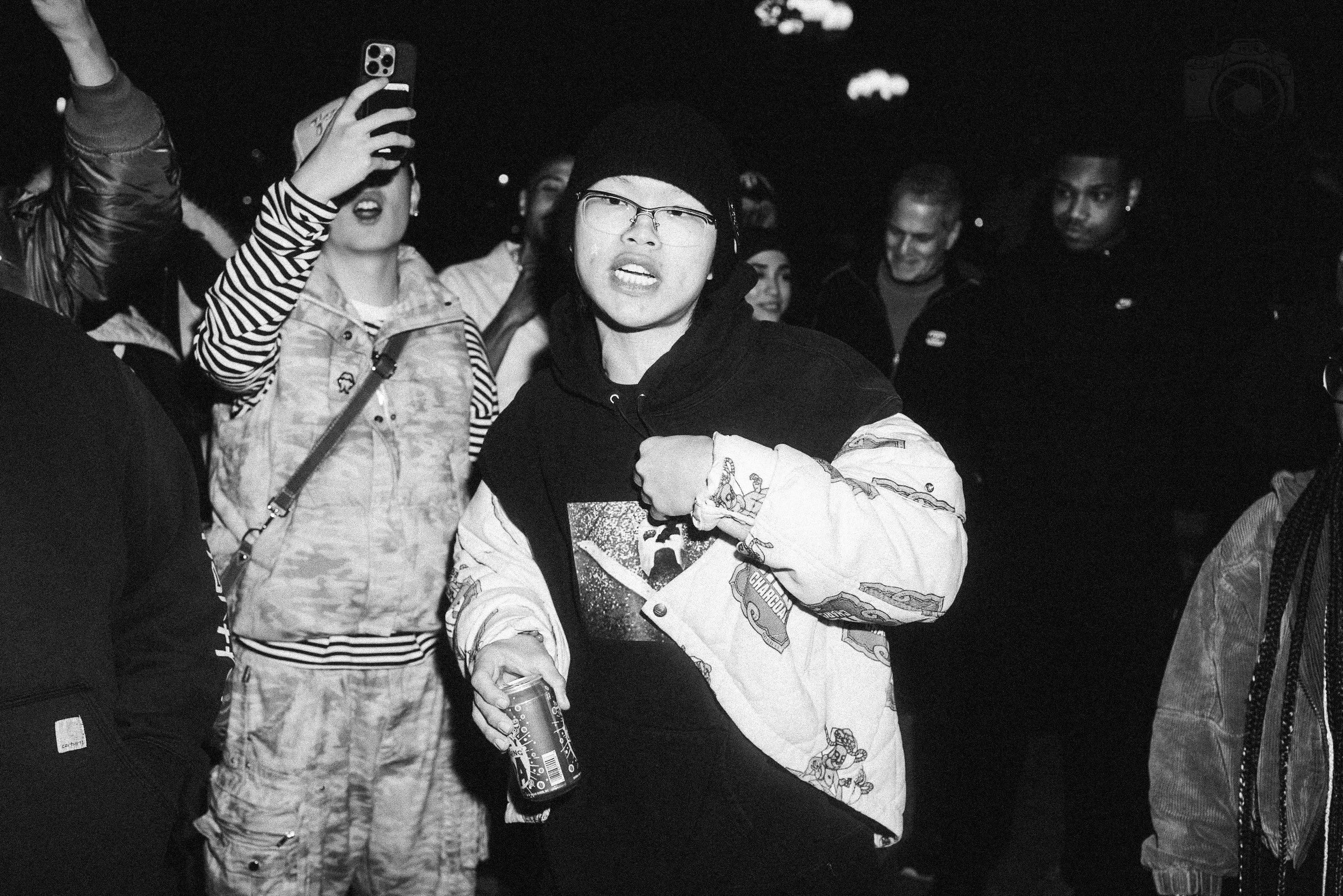 A group of young people gathered in a social setting, with one person in front holding a can and taking a selfie, some smiling and others with neutral expressions.