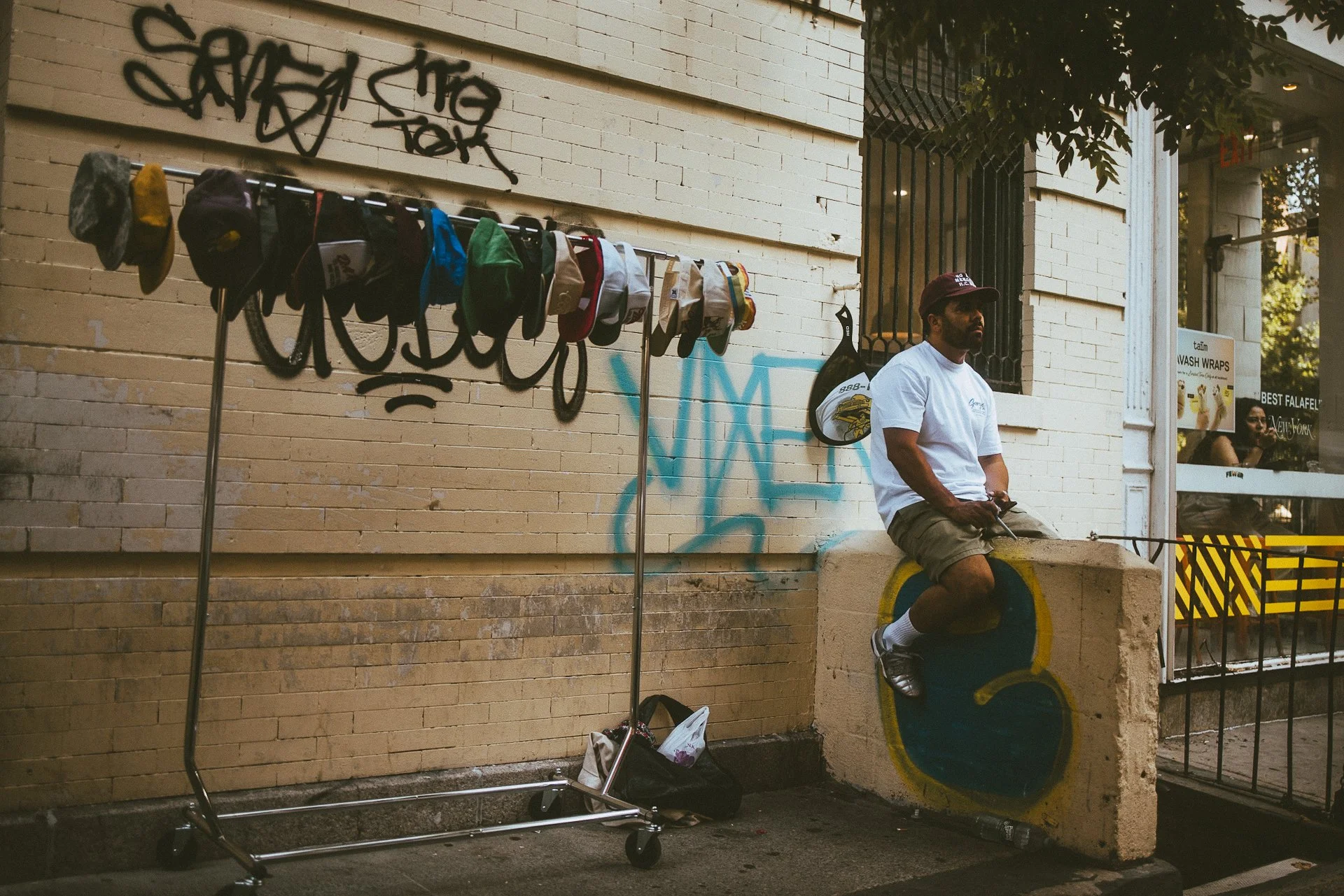 A man in a white t-shirt, khaki shorts, and a baseball cap sitting outside on a concrete barrier next to a brick building with graffiti. There is a clothing rack with hats hanging on it nearby and a black shopping bag on the ground. A sign in the bac