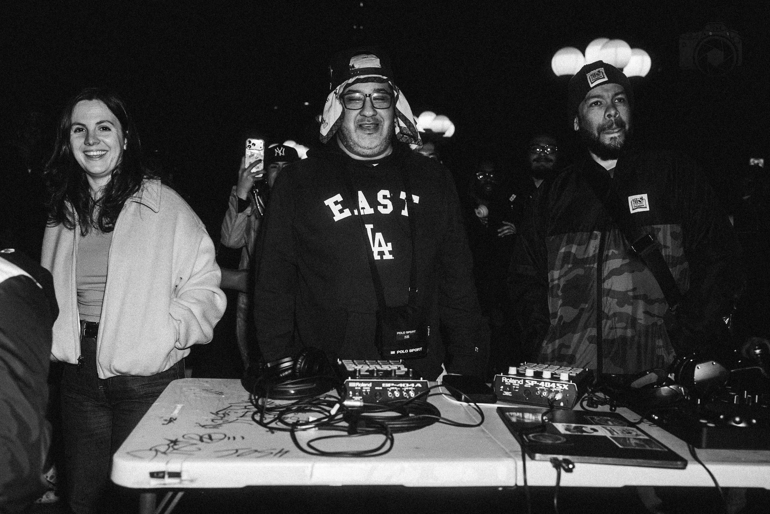 A group of people standing behind electronic music equipment at an event, including men and women smiling and dressed casually, with a dark background and hanging lights.