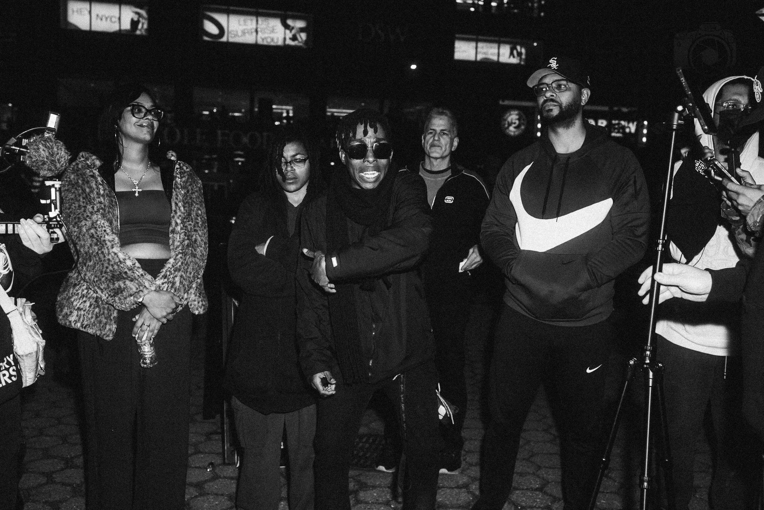 A group of people standing together at a nighttime outdoor event, with some people holding microphones and recording equipment, suggesting an interview or media coverage.