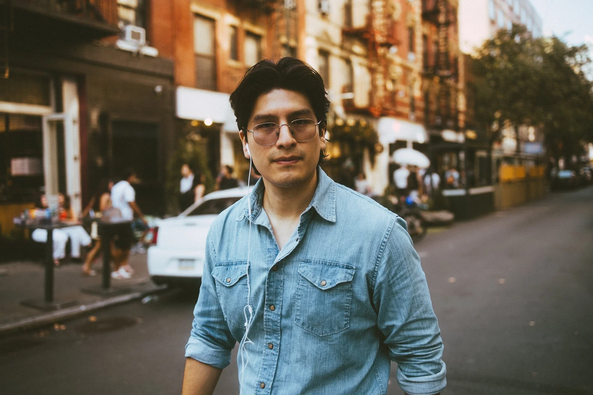 A young man with dark hair, glasses, and earphones wearing a denim shirt walking on a city street during daytime with buildings, trees, and people in the background.