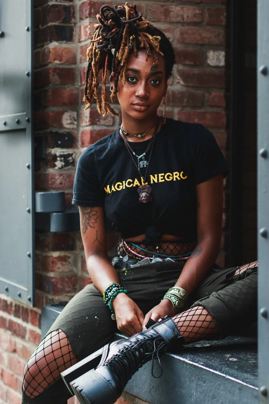 A young woman with styled dreadlocks, adorned with beads, jewelry, and a forehead ornament, sitting on a ledge in front of a brick wall. She wears a black t-shirt with the words 'MAGICAL NEGRO' and fishnet stockings under ripped pants, accessorized w