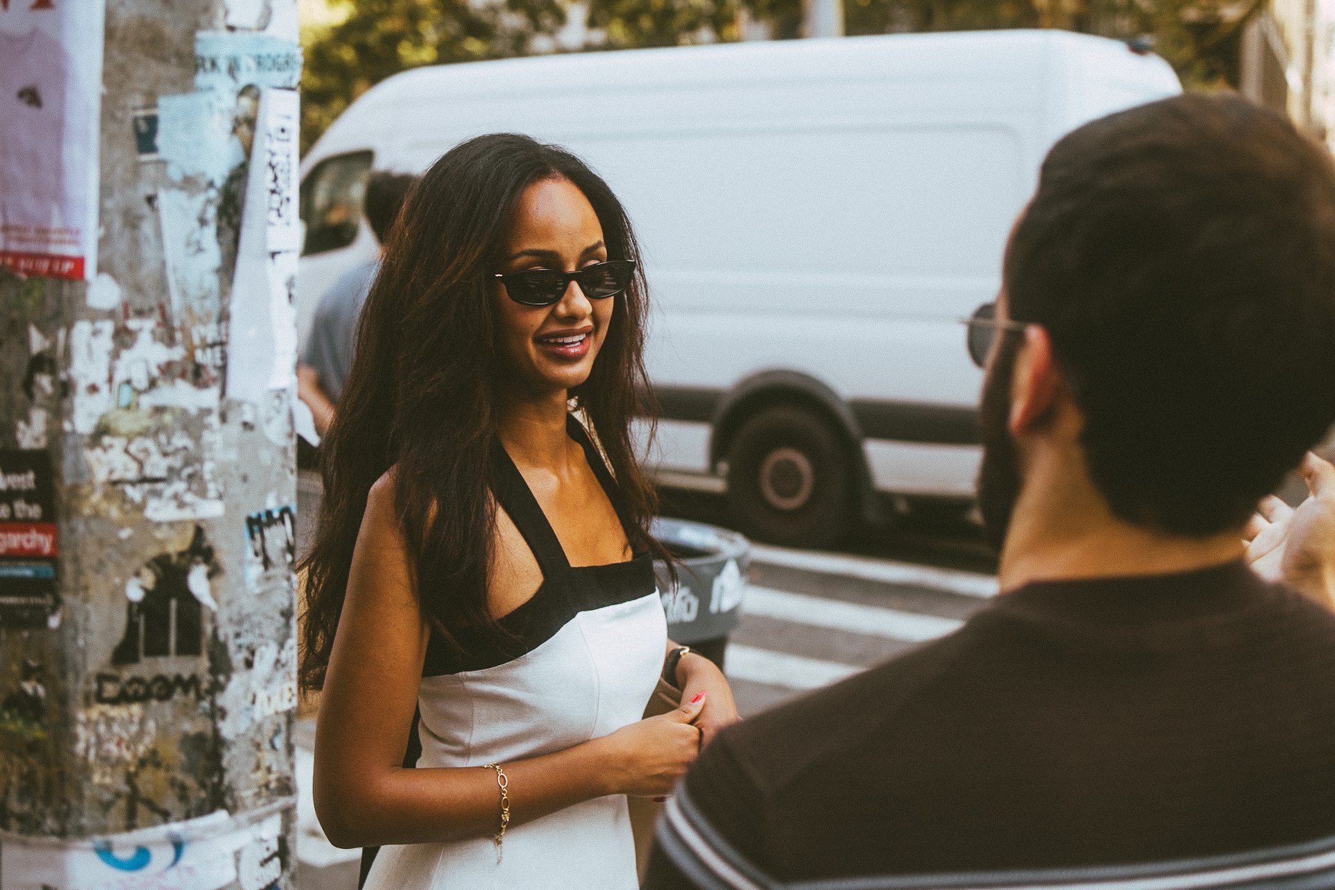 A woman with long dark hair wearing sunglasses and a sleeveless black and white dress is talking to a man with dark hair and glasses on a city sidewalk, with a white van and graffiti-covered pole in the background.