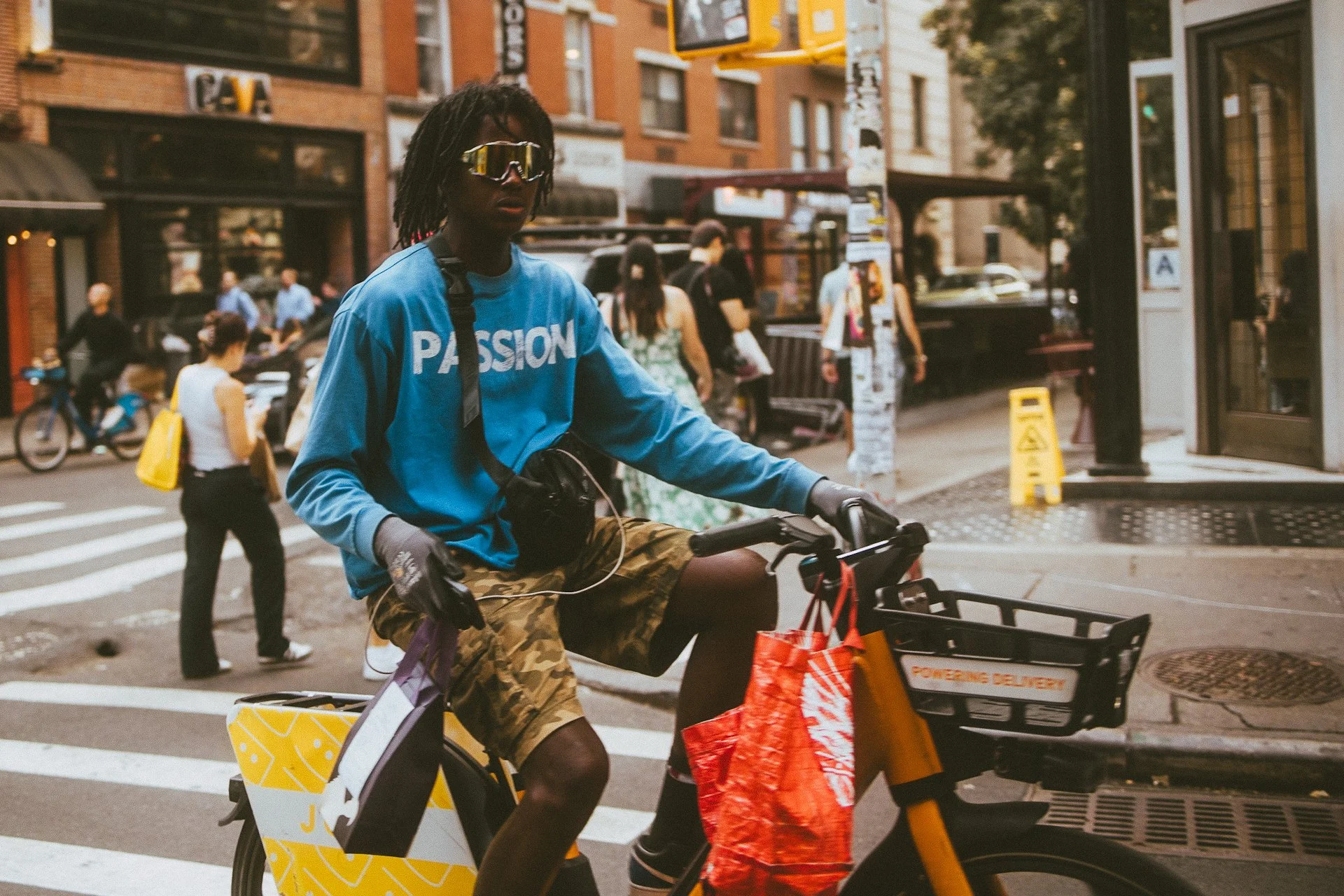 A person riding a bicycle on the street with shopping bags attached to the front. The person is wearing a blue shirt with 'PASSION' written on it, camouflage shorts, gloves, and reflective sunglasses, with a cityscape in the background.