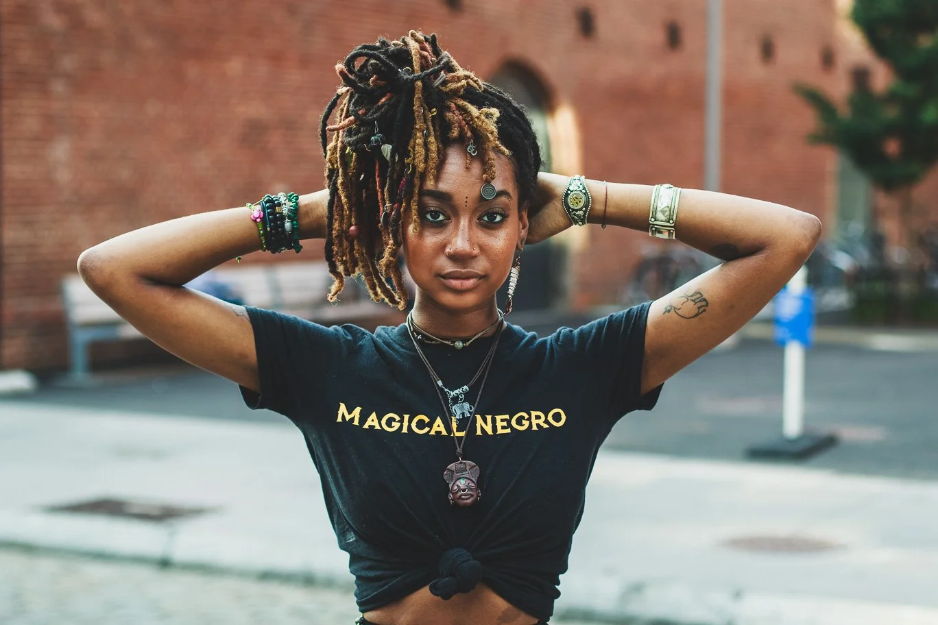 Portrait of a young woman with dreadlocks, wearing a black t-shirt with gold text, jewelry, and tattoos, standing outdoors in front of a brick building.