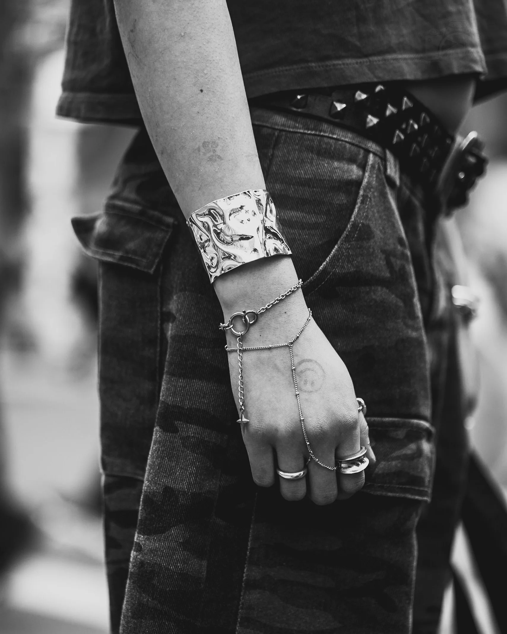 Black and white close-up of a person's arm and hand wearing layered jewelry, a leather bracelet, rings, and chains, with denim clothing.