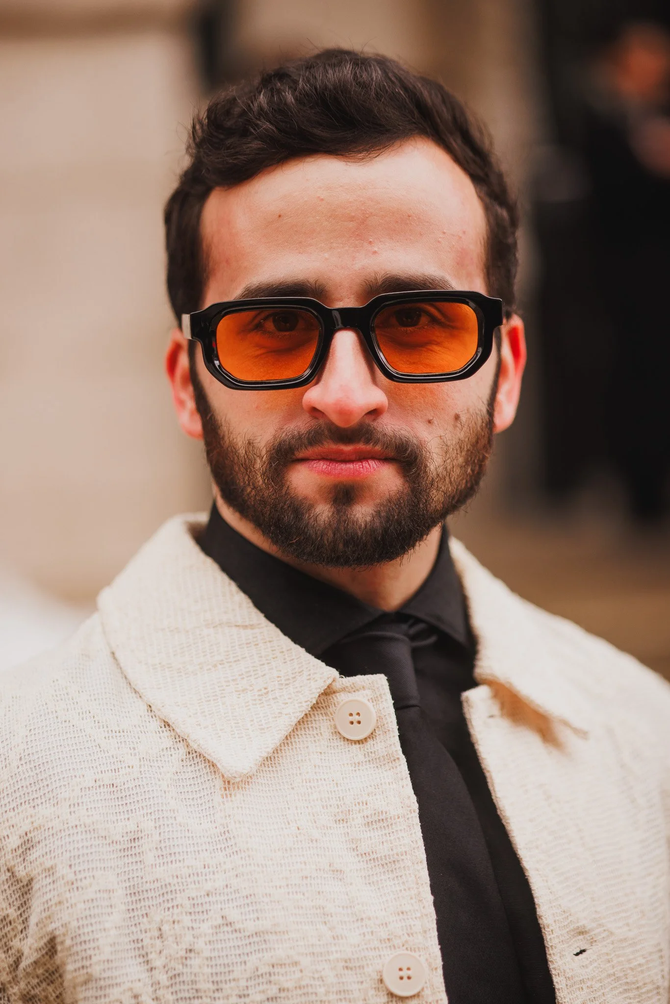 Social Media Influencer David Carmi is sporting a beard, wearing orange-tinted sunglasses, a black shirt, a black tie, and a textured cream-colored jacket.