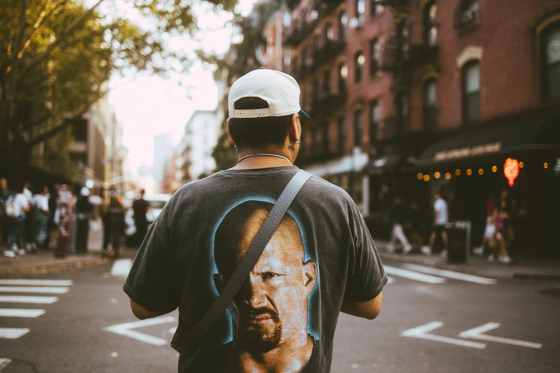Back view of a person with short hair wearing a white baseball cap, a casual t-shirt with a face graphic on it, and a shoulder bag, walking through a city street during daytime with people and brick buildings in the background.