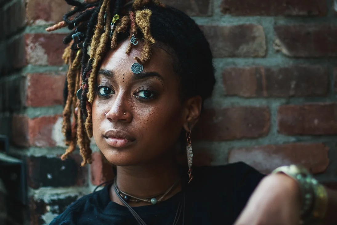 Portrait of a woman with dreadlocks styled with beads and jewelry, standing against a brick wall.